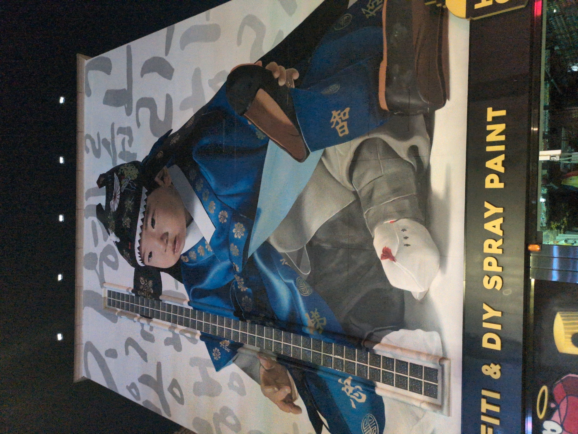 The image captures a public advertisement in Seoul, South Korea. The advertisement is a large mural featuring a young boy dressed in traditional Korean attire (hanbok). The mural depicts the boy holding a rectangular object with multiple grid-like divisions, resembling a traditional musical instrument or cultural artifact. The mural is painted on a light background with stylized, abstract gray brushstrokes. To the right of the mural is a sign that reads "FITI & DIY SPRAY PAINT" in large, bold, yellow letters. The image is taken at night, with reflections from city lights visible in the background, particularly in the signage. The scene is urban, with evidence of other nearby buildings.