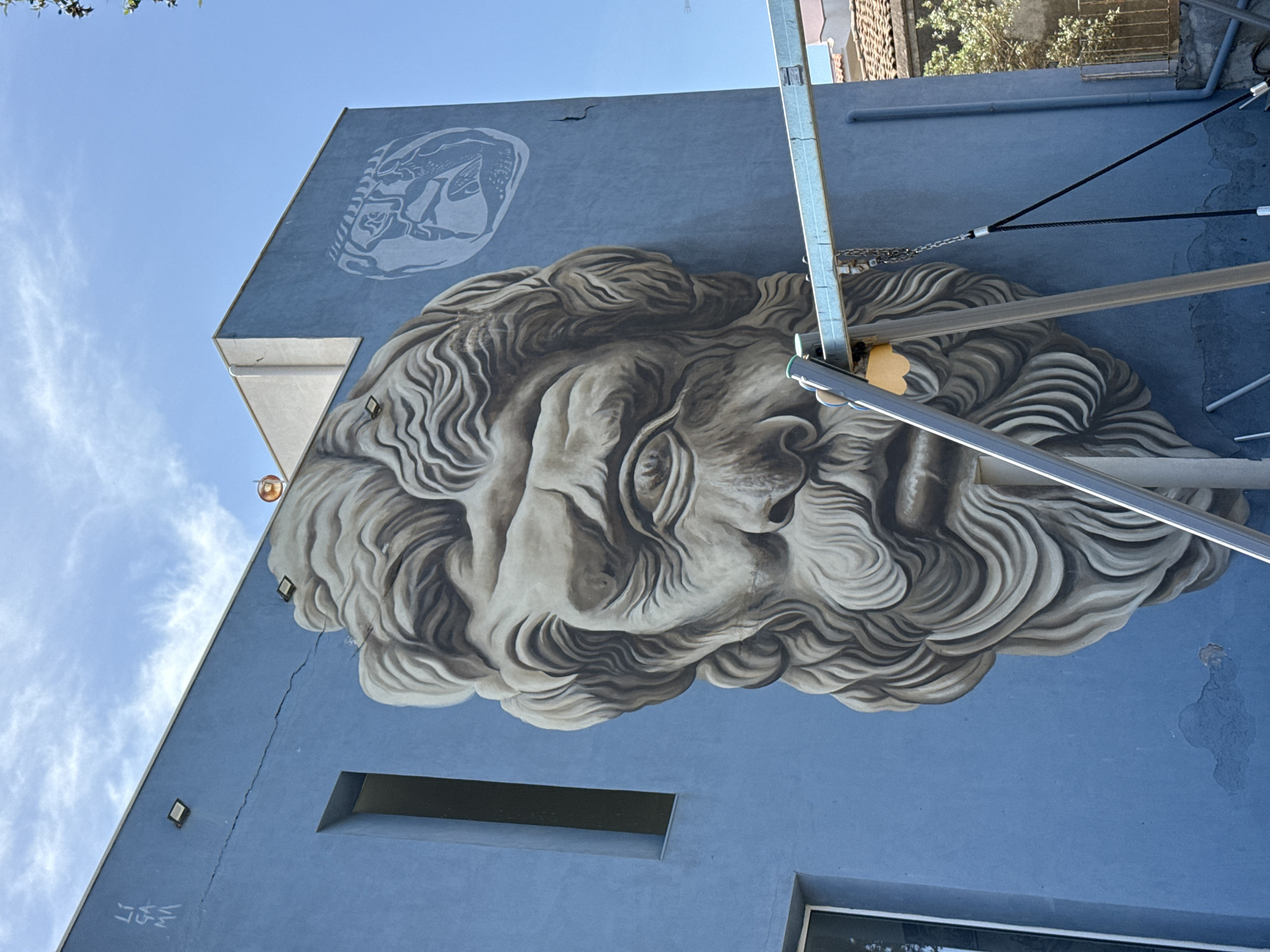 A large-scale mural adorns the side of a light blue building in an unidentified city. The principal subject of the mural is the face of a man with detailed, curly hair and a beard. Above the man's head is a lighter, smaller drawing of what appears to be a fish or sea creature. The building also features rectangular windows and attached metal supports and cables. The sky is visible above, with some clouds.