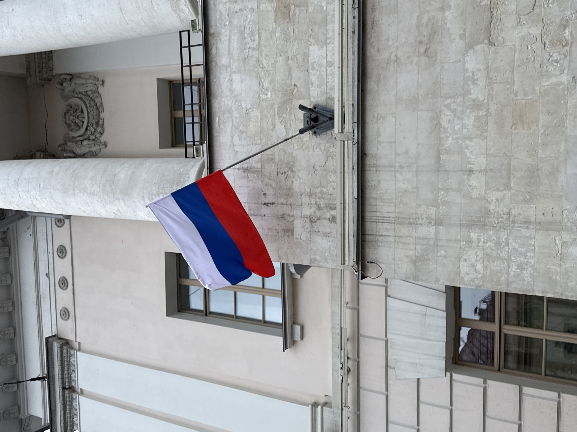 A Russian national flag, displaying horizontal stripes of white, blue, and red, is prominently flown from a flag pole extending from the facade of a building in Moscow, Russia. The image is oriented such that the building's walls appear horizontal and vertical.
The flag pole is affixed to a weathered, light grey concrete or stucco wall that shows signs of aging, with patches of peeling finish and a visible grid pattern. A triangular grey bracket secures the pole to the wall. Above and to the right of the flag (in the original orientation), a vertical metal structure, possibly a pipe or conduit, runs along the wall.
To the left of the flag, the building features more elaborate architectural details. These include large, rounded elements resembling columns or pilasters. An intricate, circular stone relief, possibly depicting a floral or classical motif, adorns a wall section between these elements. Further away, another facade, painted in a lighter, smooth white, exhibits decorative horizontal cornices and smaller, circular rosettes.
Several windows are visible across the different sections of the building. One window directly below the flag has a brown frame and reflects part of the surrounding environment, including what appears to be another building or roof structure. Another window with a dark frame is visible in a deeper recess of the building's facade.
The lighting is soft and diffused, suggesting an overcast day with no harsh shadows. There is no visible text. The overall scene depicts a grand, possibly historic or official, building displaying the national flag.