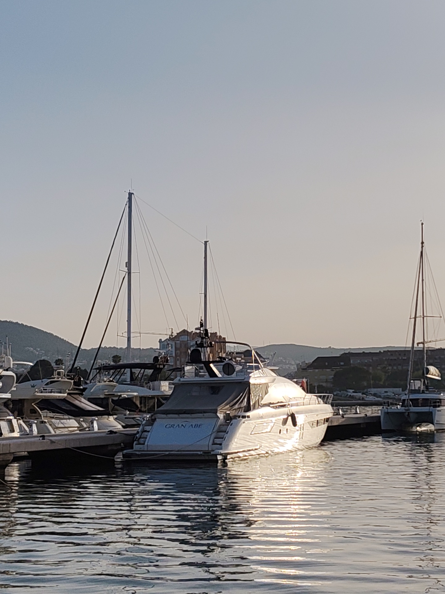 The image shows several yachts docked in the marina of Dénia, Spain. The water is calm, reflecting the sunlight. A yacht in the center of the frame displays the name "GRAN ABE" on its stern. The marina is situated with buildings and hills in the background under a clear sky.