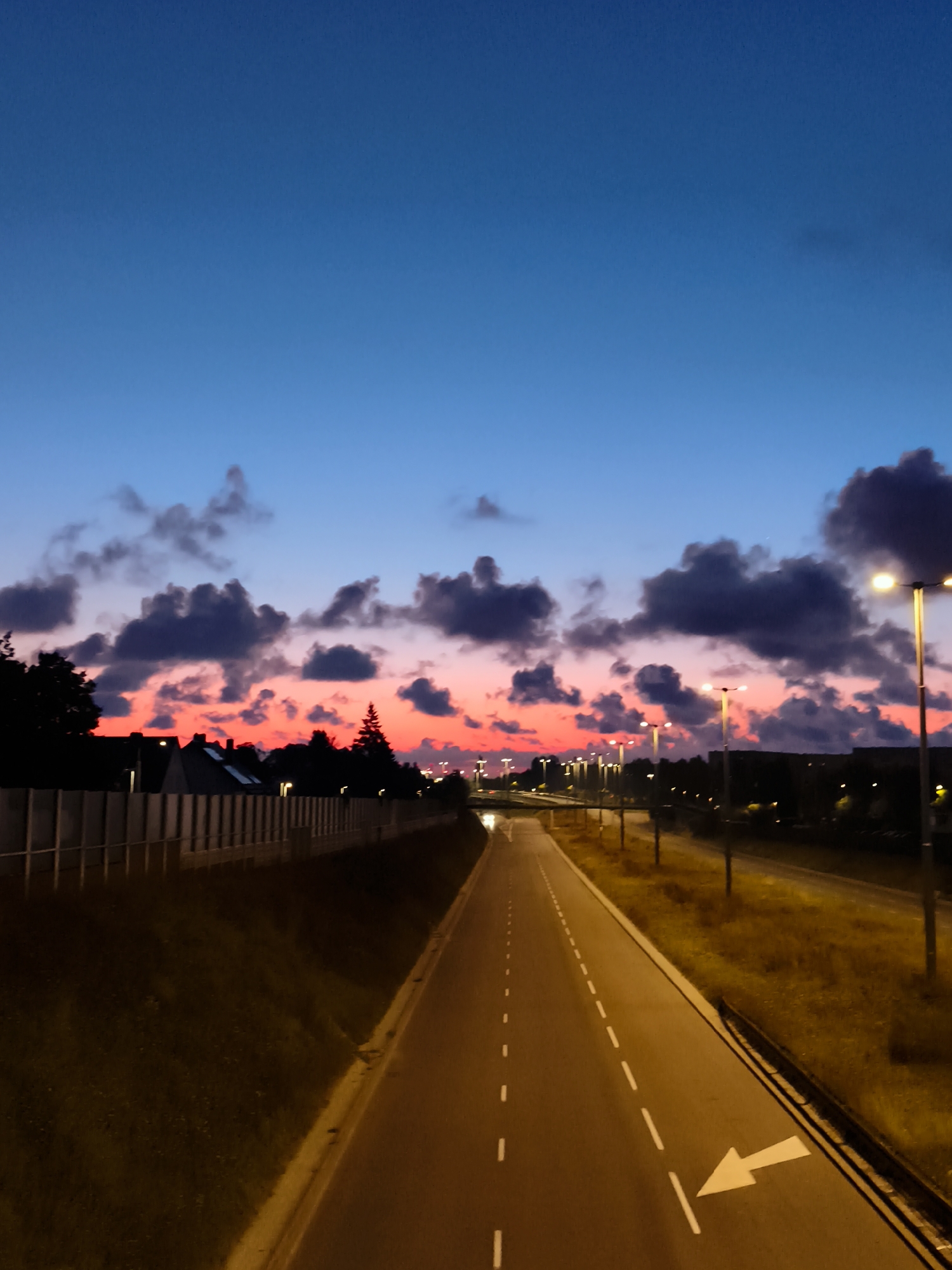 A long, empty road with streetlights lines a path through a suburban area at dusk. The sky above is a gradient of deep blue to a vibrant pink and orange, with dark clouds scattered across the horizon. The road is paved with asphalt and marked with white dashed lines. On the right side of the road, there is a grassy embankment with sparse vegetation. On the left side, a concrete barrier runs along the edge of the road, with silhouetted houses and trees visible behind it. The scene appears to be located in Gdańsk, Poland.