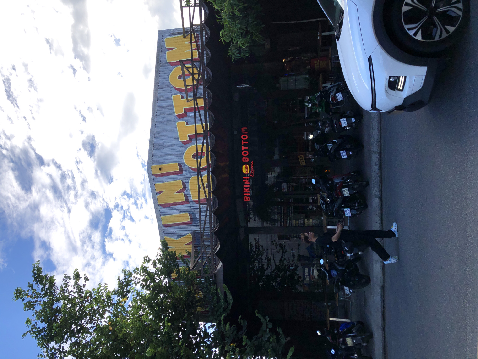 The image captures a street scene in Đà Nẵng, Vietnam, in front of a restaurant called "Bikini Bottom Express." The restaurant has a distinctive facade with large, yellow, three-dimensional lettering spelling out "BIKINI BOTTOM" against a corrugated metal background. Below this, a red neon sign displays "BIKINI BOTTOM Express" with a burger graphic. A person, dressed in black pants and a black t-shirt, walks along the sidewalk in front of the restaurant. They are wearing white and light blue footwear and have earbuds in. To the left of the person, several motorbikes are parked along the curb. On the right side of the frame, the front of a white car is visible, parked on the street. The sky above is partly cloudy, with a mix of blue sky and white clouds, and a large green tree with dark green leaves is present on the left side of the image. The overall setting suggests an urban commercial area.