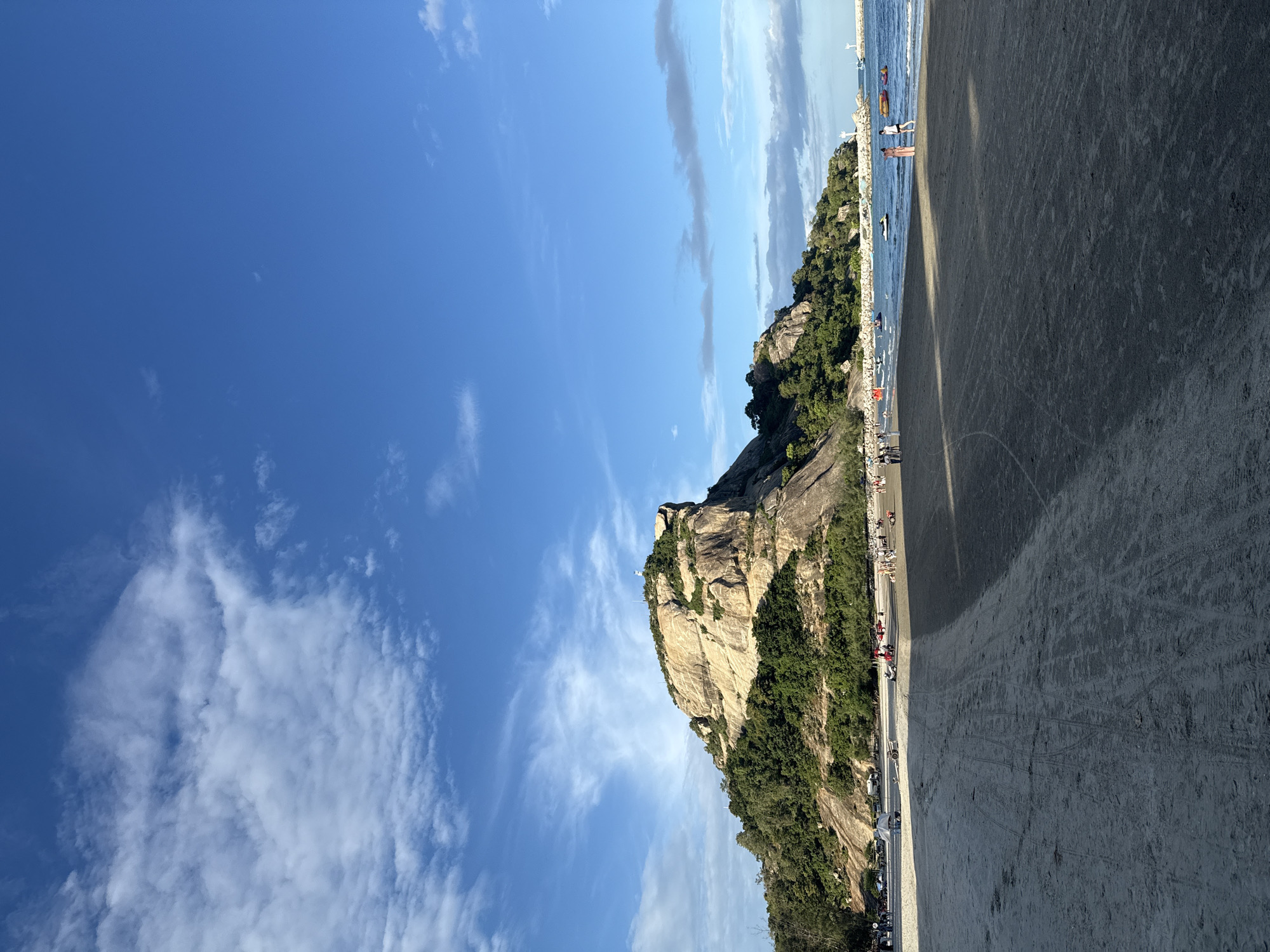 The image depicts a panoramic view of Hua Hin Beach in Hua Hin City Municipality, Thailand. A large, rocky hill covered with lush green trees dominates the background, with a prominent rock formation at its summit. The foreground is a sandy beach with tire tracks etched into the sand, indicating vehicular or vehicle activity. The ocean stretches out towards the horizon, with several small boats and jet skis visible on the water. A lighthouse stands on a pier or breakwater in the distance. Several people are scattered across the beach, some walking, others seemingly relaxing or engaging in activities near the water. The sky is a clear blue with scattered white clouds, suggesting pleasant weather. The overall scene captures a typical beach day with natural beauty and recreational opportunities.