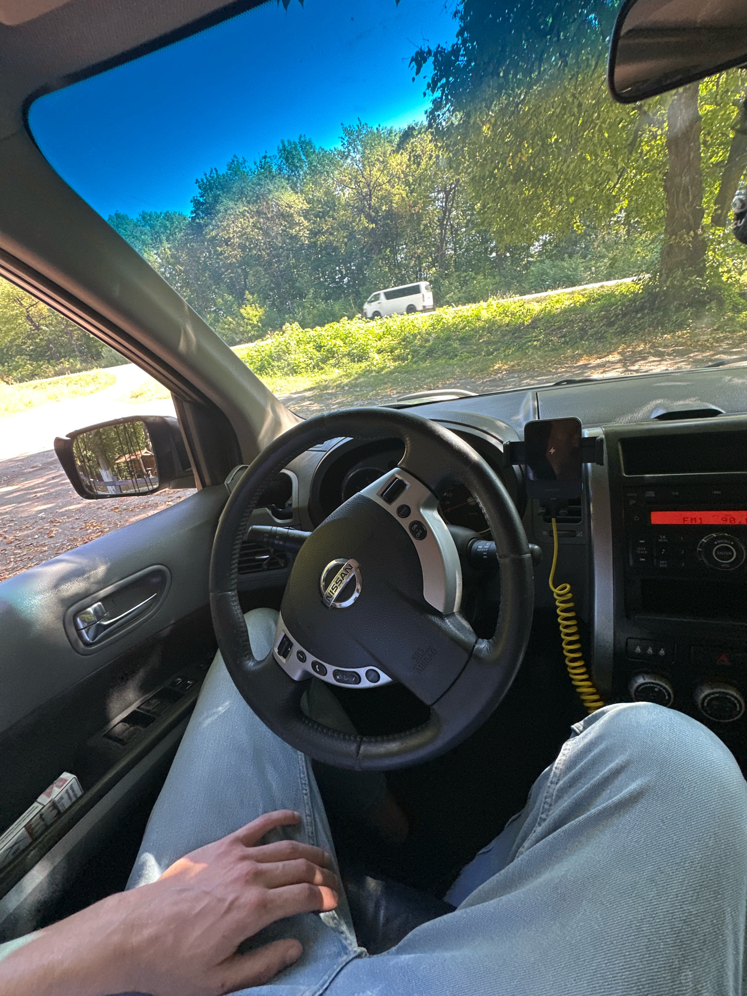 The image provides an interior view from the driver's seat of a Nissan vehicle, parked on a dirt road. The driver's left hand rests on their lap, which is covered by light blue jeans. The steering wheel, with the Nissan logo visible, is in the foreground. A smartphone is mounted on the air vent to the right of the steering wheel. The dashboard displays a radio with "FM 1 90.0" visible on the screen. Through the windshield, a white van is seen driving on a paved road surrounded by lush green trees and foliage. The sky is a clear, vibrant blue. A side-view mirror reflects trees and what appears to be a structure, possibly a gazebo or cabin. The context of Shatava, Ukraine is not discernible from the visual information present in the image.