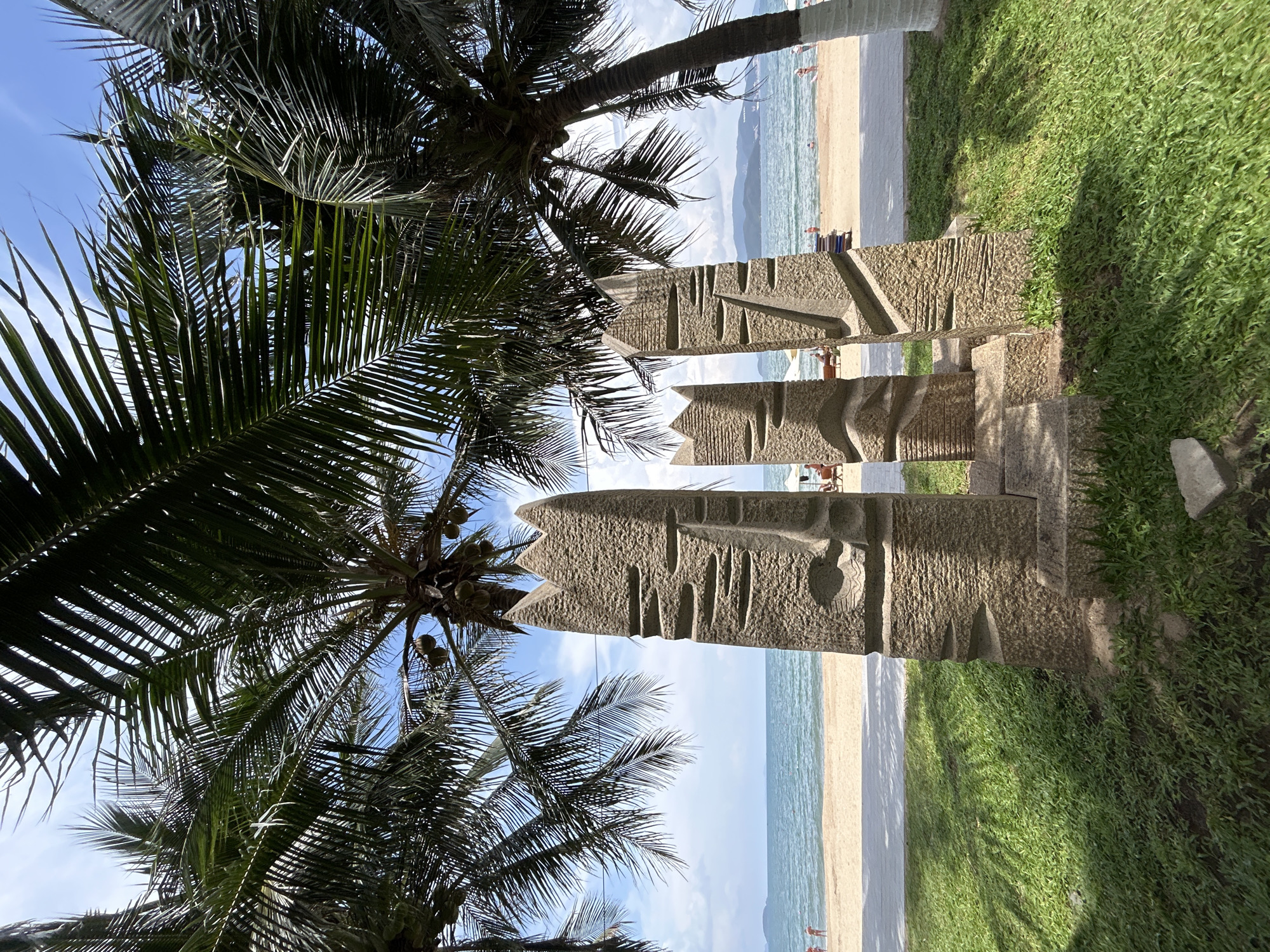The image shows a beach scene in Nha Trang Ward, Vietnam. In the foreground, there are three carved stone pillars with abstract facial features standing on a grassy area. Palm trees are positioned on both sides of the pillars, partially obscuring the sky. The beach is visible in the background with sand, water, and distant figures of people. The sky is blue with some clouds.