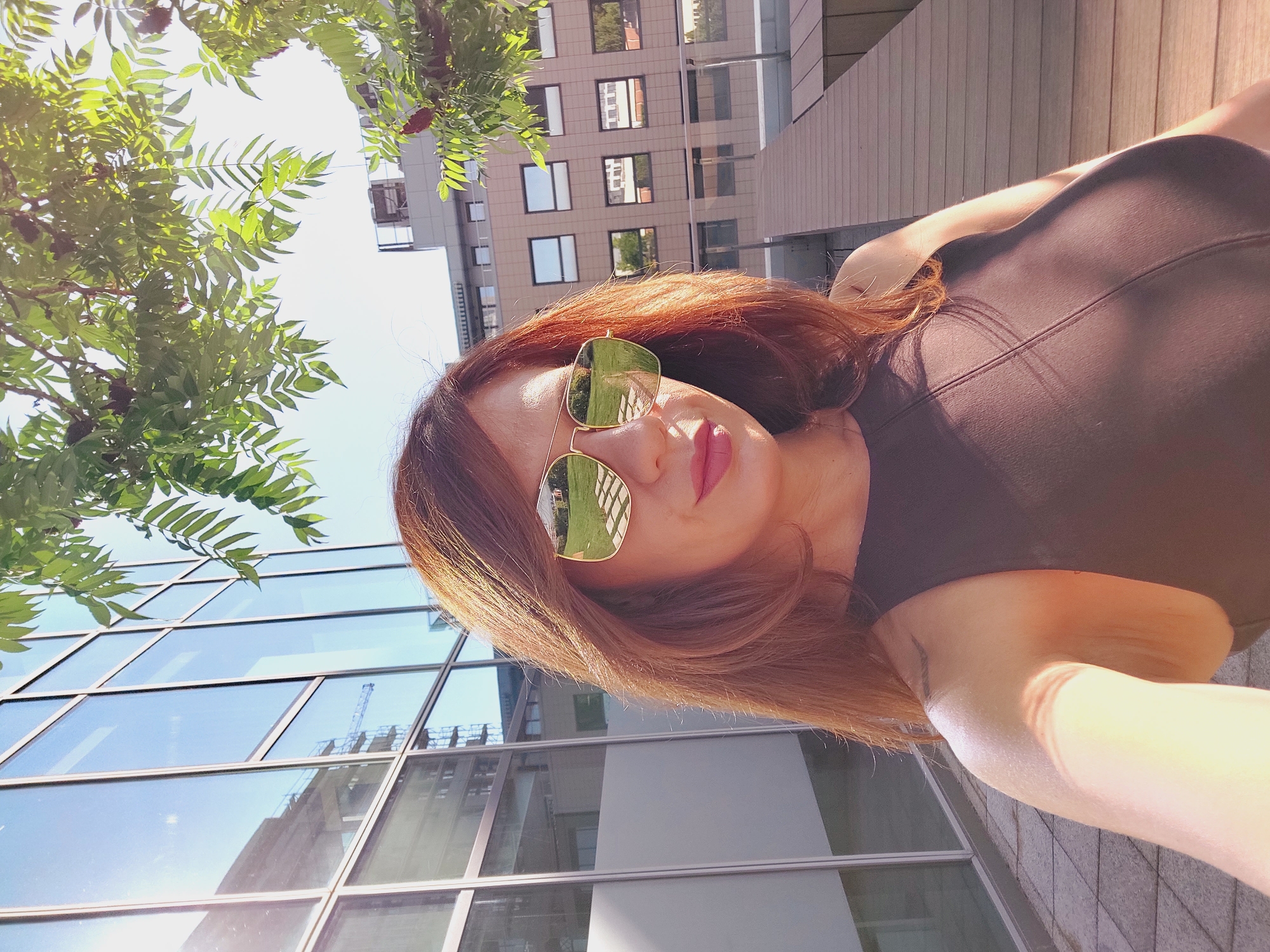 A woman with auburn hair and wearing sunglasses and a dark sleeveless top is taking a selfie. She is standing outdoors in front of a modern glass-fronted building, with a tree in the foreground. The sunglasses reflect a green grassy area with a paved walkway. In the background, another building with many windows is visible, along with some wooden decking. The scene appears to be in an urban environment, possibly a city. The sunlight is bright, casting shadows and highlighting parts of the woman's arm and shoulder. A faint tattoo is visible on her upper left arm. The location is the city of Sofia, Bulgaria.