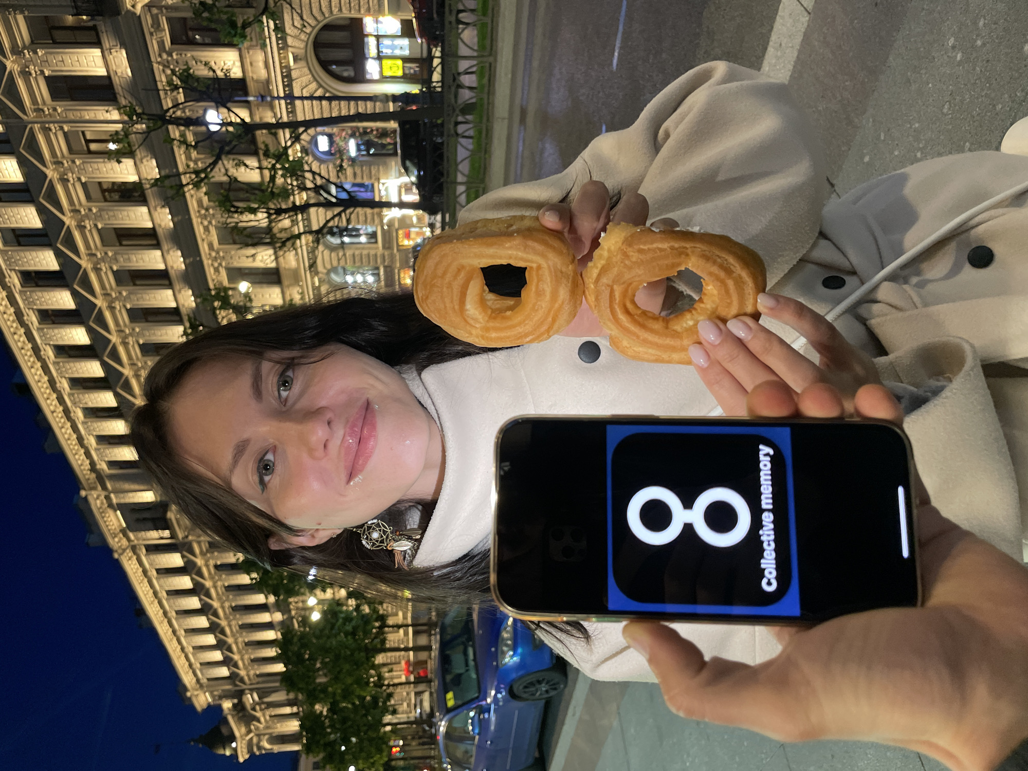 A woman stands in front of a well-lit building in Saint Petersburg, Russia, at dusk or night. She is holding two pastries that appear to be choux rings. In her left hand, she holds a smartphone displaying a screen with a white number "8" and the words "Collective memory" below it. The building behind her is ornate and illuminated, with multiple windows. A blue car is parked on the street to the left, and trees line the sidewalk. The woman is wearing a light-colored coat and a distinctive earring. The overall scene suggests a casual outing in an urban environment.