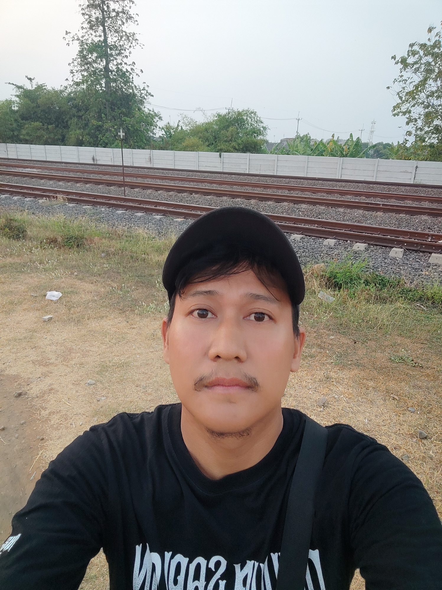 A man in a black t-shirt and black baseball cap is taking a selfie outdoors. He has short, dark hair and a mustache. He is standing in front of a set of train tracks. Behind the tracks, there is a concrete wall and some trees. The sky is overcast. The location is likely Pakusamben, Indonesia, given the presence of train tracks and tropical vegetation.