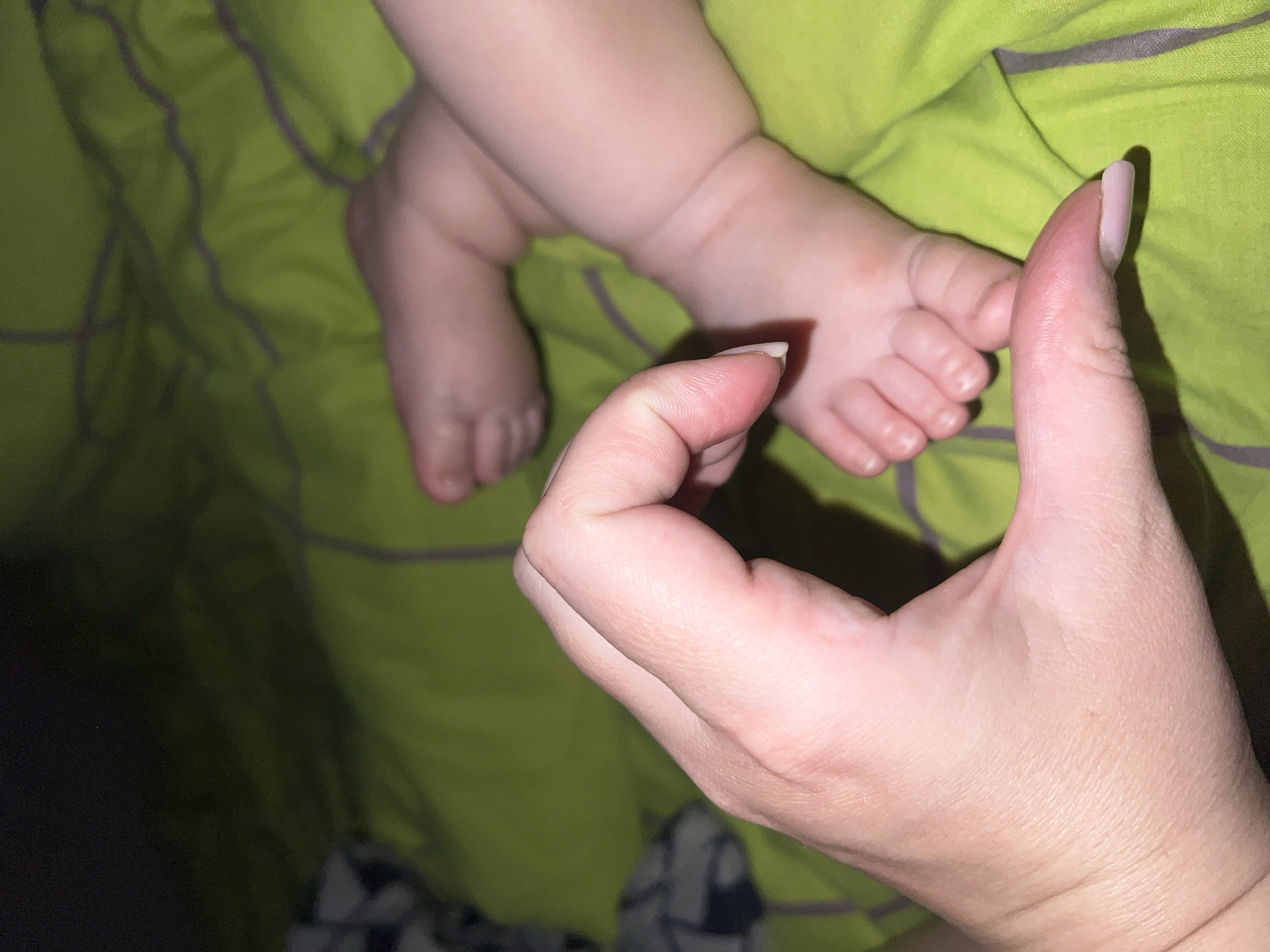 A close-up shot shows a person's hand forming a heart shape with their thumb and index finger, positioned over a baby's feet. The baby's feet are resting on a green blanket with gray stripes. The hand is in the foreground, with the fingers gently curved. The baby's feet are slightly out of focus in the background. The lighting is dim, creating a soft, intimate atmosphere. The image does not provide any information about the location, as it is not specified.