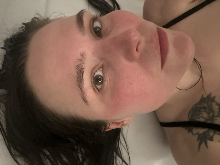 The scene is located in Bartula, Montenegro. The image presents a close-up, angled view of an adult female, appearing to lie down or recline, gazing directly at the camera. Her face occupies the majority of the frame, with the top of her head angled towards the upper left. She has dark, medium-length hair, visible around her forehead and sides, appearing slightly damp or disheveled. Her skin tone is light, with a noticeable pink flush across her cheeks and nose. Her eyes are brown, with distinct circular light reflections visible in both pupils, indicative of a ring light source. Her eyebrows are dark and moderately thick. Her lips are full with a neutral to slight upturned expression. The upper part of her left shoulder and collarbone are visible in the lower right, displaying a black strap, likely from a tank top or similar garment. A tattoo, featuring a floral design resembling a peony, is visible on her left shoulder. The background is a plain, undifferentiated light-colored surface, possibly white or off-white, suggesting an indoor environment such as a bathtub or bed.
