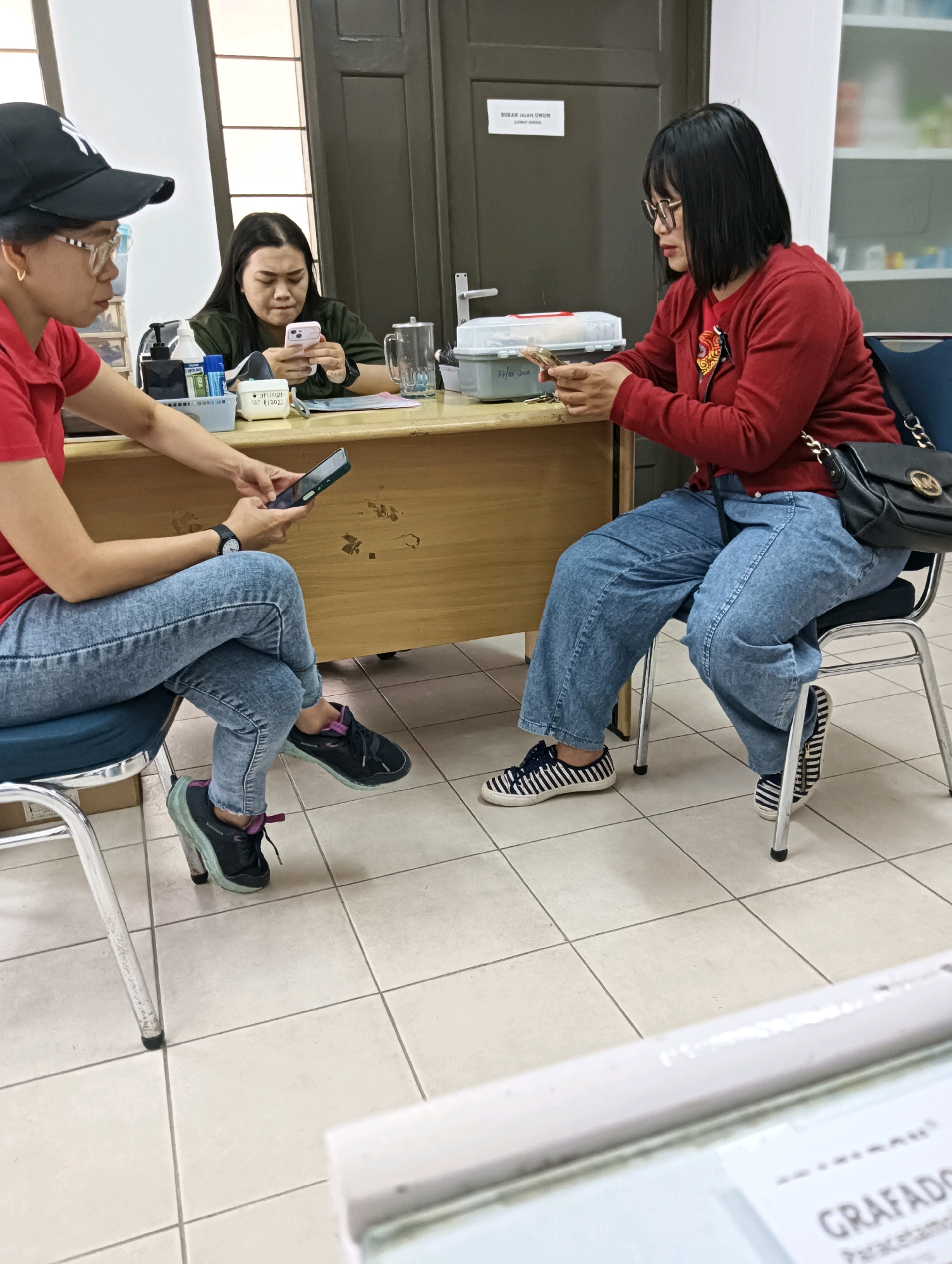 In Tomohon, Indonesia, three individuals are seated at a wooden counter. The person on the left, wearing a red shirt and a black baseball cap, is looking at their phone. The person in the middle, in a green shirt, is also looking at their phone. The person on the right, in a red cardigan and blue jeans, is also looking at their phone. Various items are present on the counter, including a tissue box, a container, and a pitcher. The scene appears to be an indoor waiting area or office space.