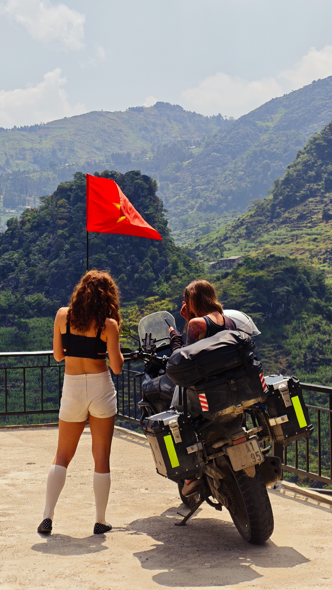 Two women stand near a parked touring motorcycle on a concrete platform with a black metal railing. The motorcycle is heavily laden with luggage and panniers. A red flag with a yellow star flies from a pole nearby. The woman on the left has red curly hair, wears a black top, white shorts, and knee-high socks with patterned shoes. The woman on the right has brown hair, a tattoo on her arm, and is looking at a phone. The motorcycle's license plate is visible. Behind them is a mountainous landscape covered in dense green vegetation, under a sky with some clouds. The scene is likely in Lũng Phìn Commune, Vietnam.