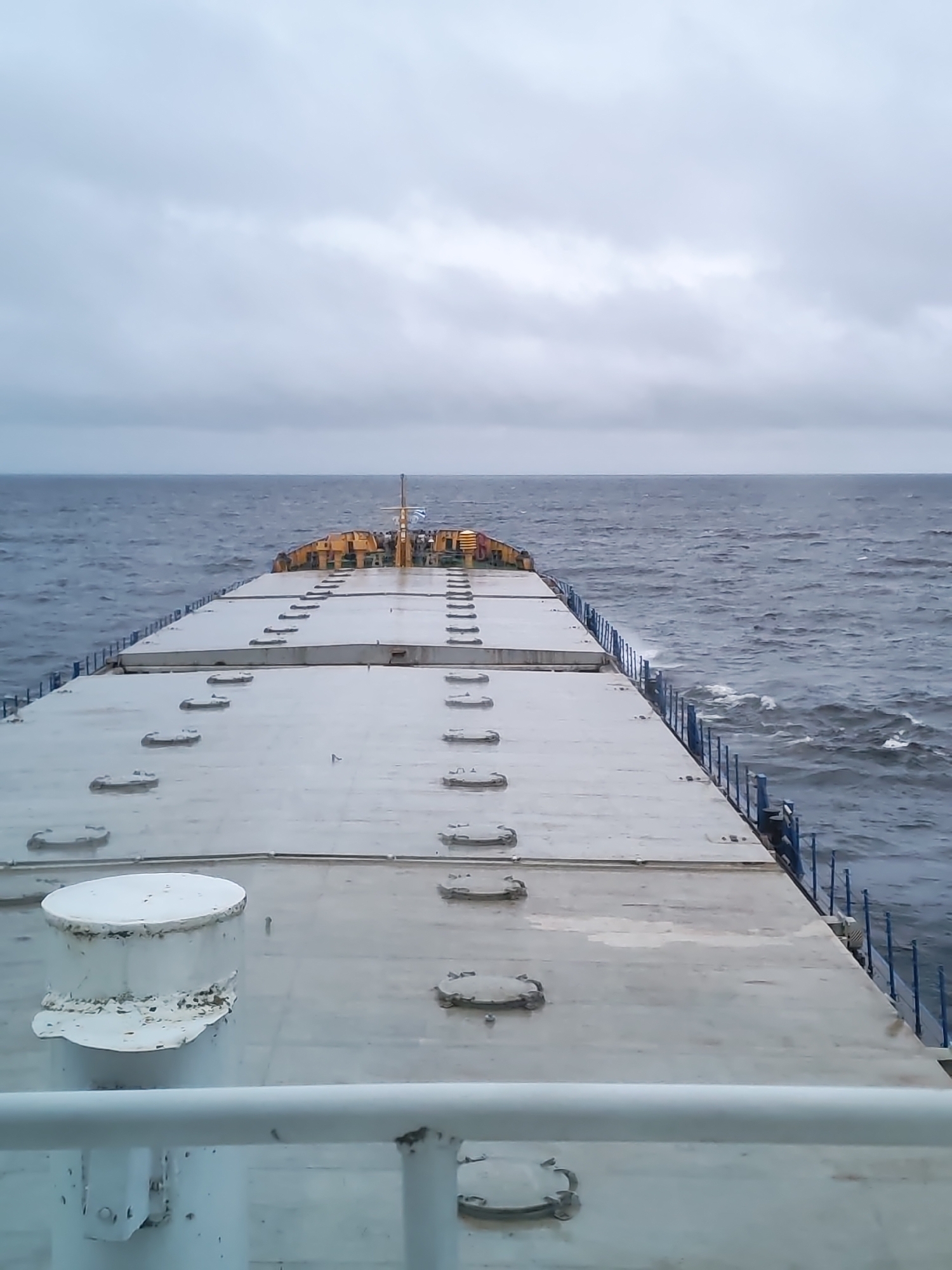 The image depicts the deck of a ship at sea. The deck is gray and has several hatches. In the foreground, there is a white bollard and a white railing. The sea is choppy and gray. The sky is overcast with dark, gray clouds. There are no people visible. The location is likely not in the city of Ozernoe Ust'e, Russia, as the city is located inland and does not have direct access to a large body of water suitable for a ship of this size.