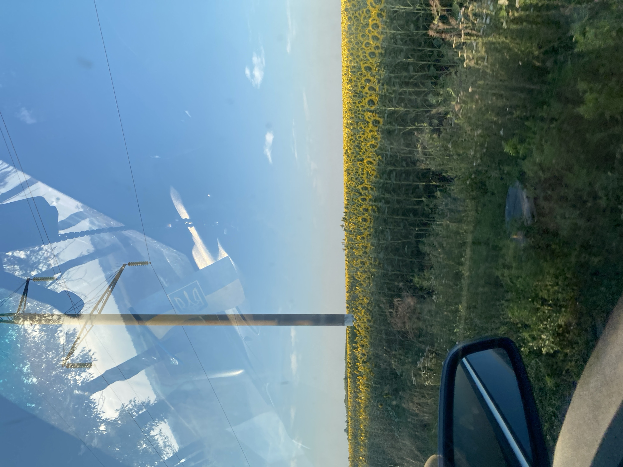 This image captures a scenic view from a vehicle in Maiske, Ukraine. A vast field of sunflowers stretches across the horizon under a clear blue sky dotted with sparse clouds. In the foreground, the side mirror of the vehicle is visible, reflecting the surrounding environment. A tall utility pole stands prominently in the middle ground, with power lines extending across the frame. The reflections on the vehicle's window add a layer of complexity, showing trees and a symbol that appears to be the Ukrainian trident. The overall impression is one of rural beauty and vast open spaces, typical of the Ukrainian countryside.