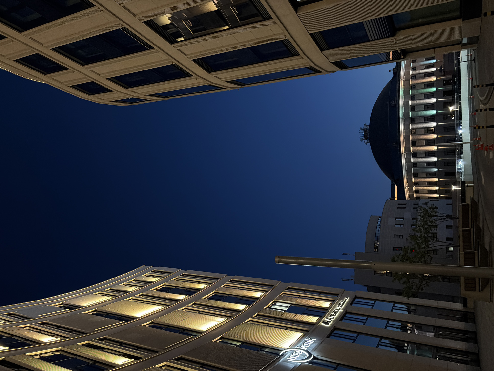 The image captures a nighttime view of the modern architectural ensemble in Minsk, Belarus, specifically focusing on the buildings surrounding the National Library of Belarus. On the left, a curved building with glowing vertical lights and signage for "neo bank" and "Альфа-Банк" is prominently featured. To the right, another curved building with dark windows and light-colored facade frames the view. In the center background, the distinctive dome of the National Library of Belarus is illuminated with multi-colored lights, standing out against the deep blue night sky. The foreground shows a paved area with benches, trees, and safety cones, indicating a public space. The overall scene conveys a sense of urban development and modern design under the night sky.