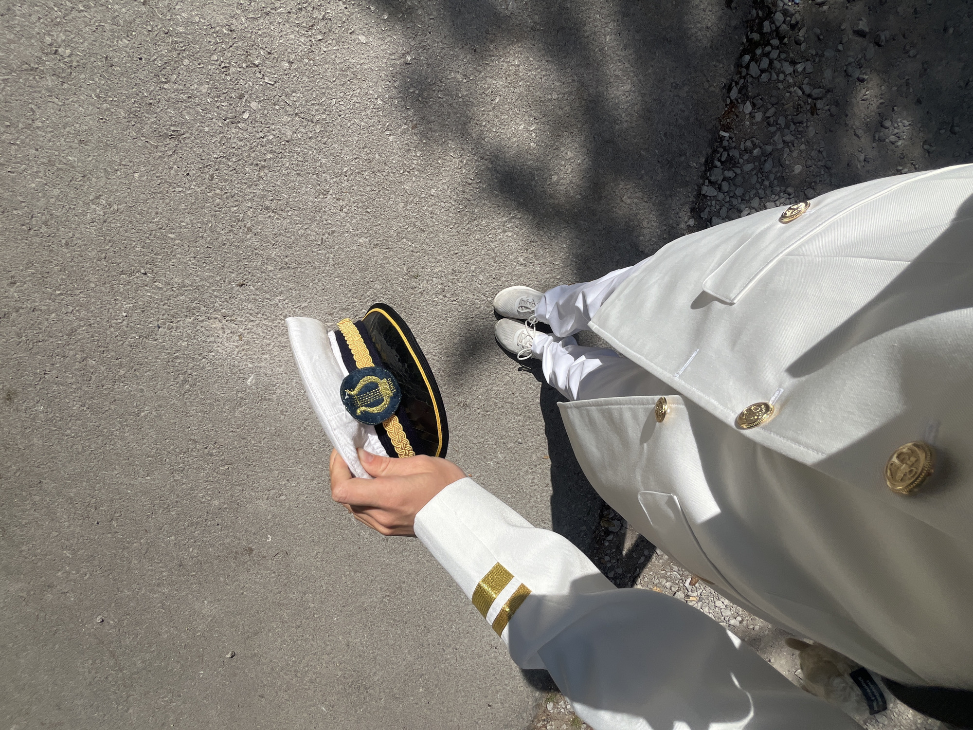 A high-angle, first-person perspective shows a person in a white military uniform holding a white peaked cap trimmed with gold braid in their left hand. The cap has a round emblem on the front. The uniform is white with gold buttons and gold stripes on the sleeve cuffs. The person is wearing gray sneakers. The person is standing on an asphalt surface with shadows of tree branches. This image shows no information that allows identification of the location in Budva, Montenegro.
