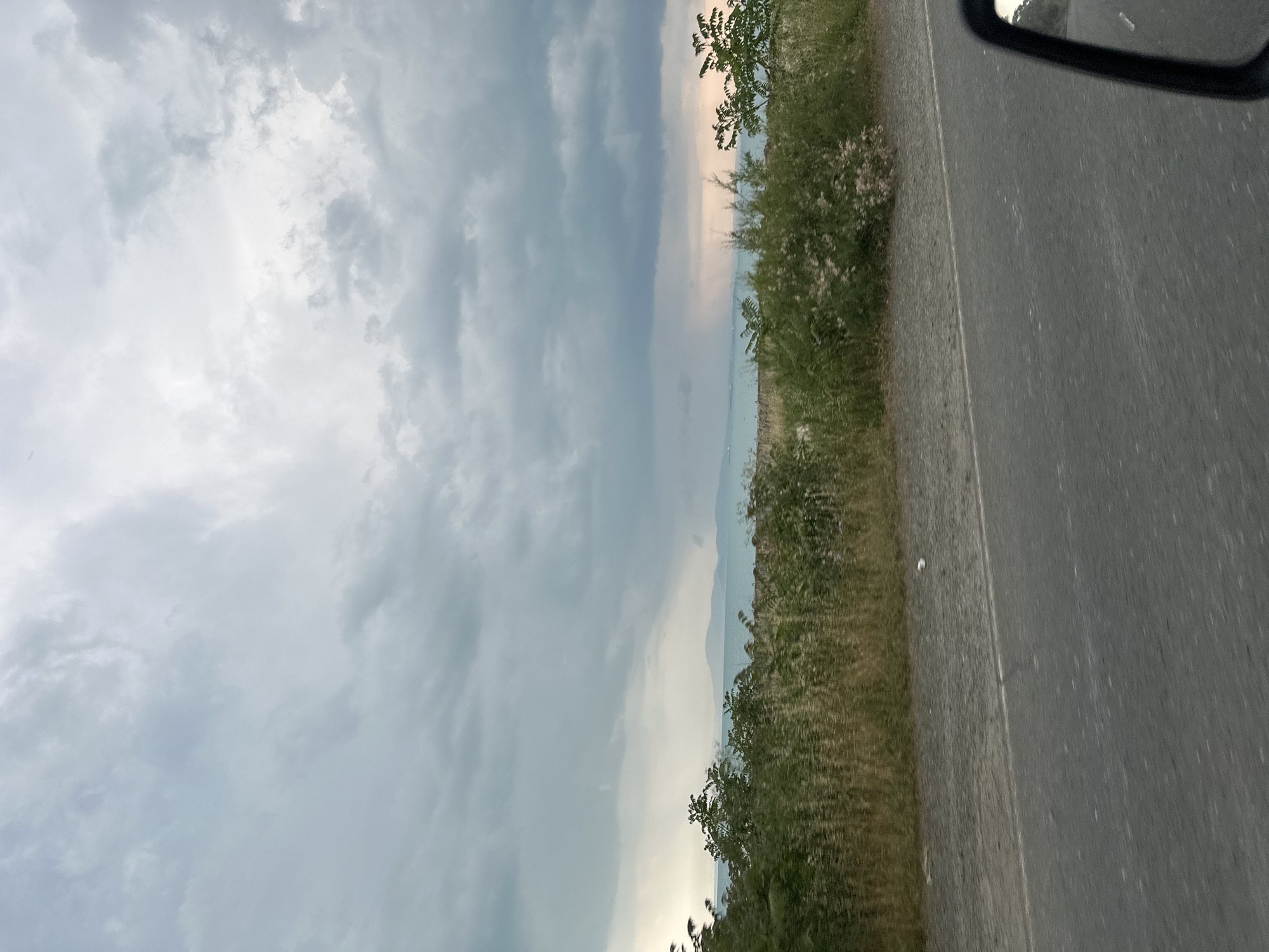 The image, correctly oriented horizontally, captures a landscape view from the perspective of a moving or stationary vehicle. On the far right, a dark gray asphalt road with a faint white dividing line is visible, suggesting a highway or rural route. Immediately to the left of the road is a strip of dense, natural vegetation, comprising various green and brownish bushes, grasses, and some scattered trees, consistent with a roadside or embankment. Beyond this foliage, a vast, hazy horizon extends, featuring the indistinct outlines of distant landforms, possibly hills or mountains, and a subtle suggestion of a large body of water beneath the cloud cover. The majority of the left side of the frame is filled with an overcast sky, characterized by varying shades of gray and white clouds. There are some brighter patches in the sky, hinting at sunlight attempting to penetrate the cloud cover. A small part of a vehicle's black side-view mirror frame is visible in the upper right corner. The scene appears to be set during daytime, likely late afternoon, with diffused lighting due to the heavy cloud cover. No people or discernible text are present. The overall impression is a serene, cloudy day while traveling through a natural landscape.