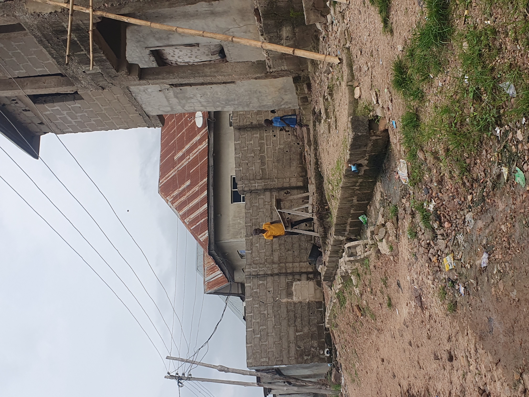 The image shows a street scene in Lokoja, Nigeria. In the foreground, the ground is covered with dirt, rocks, and debris, with some patches of green grass. A broken concrete drainage channel runs along the right side of the frame. In the middle ground, a wall constructed of grey cinder blocks separates a more built-up area from the street. Two young people are visible near this wall. A young man in a yellow t-shirt and dark pants stands near a wooden sawhorse, looking towards the camera. To his right, a younger child in a blue dress is walking away from the camera. Behind the wall, several structures are visible. One building has a weathered, reddish-brown corrugated metal roof. Adjacent to it, another building is under construction, with exposed cinder block walls and concrete supports. Electrical wires are strung across the sky, supported by wooden poles. The sky is overcast. The overall impression is one of an unpaved, developing urban area with ongoing construction and a need for improved waste management.