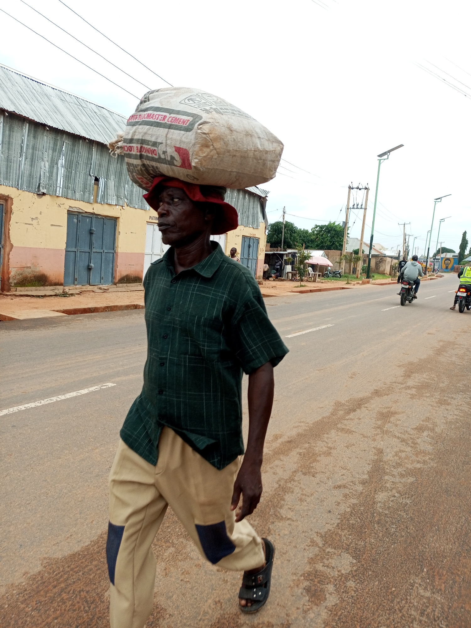A man wearing a green plaid shirt, tan pants with dark blue patches, and a red hat walks on a paved road in Tempure, Nigeria, carrying a large bag of cement on his head. The road has a white dividing line and is lined with buildings and trees. Power lines and streetlights are visible in the background. Two motorcyclists are also present on the road.