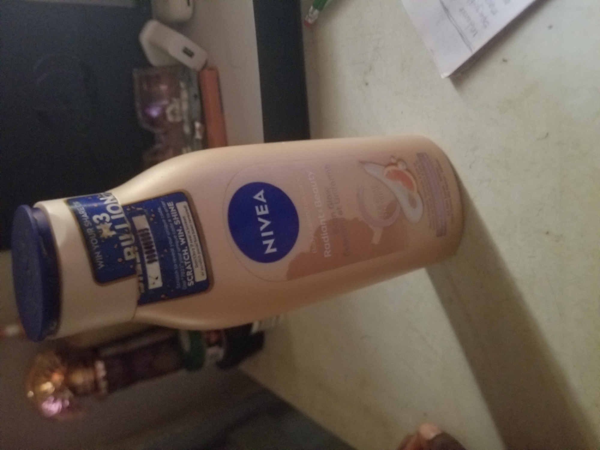 The image shows a bottle of Nivea Radiant Glow body lotion. The bottle is light peach colored with a blue cap. A blue and white label is attached to the bottle, featuring text such as "WIN YOUR SHARES", "$3 BILLION", "SCRATCH, WIN, SHINE", and "NIVEA". Below this, the text "Body Lotion", "Radiant Glow", and "Peau Radieuse et Uniforme" is visible. The background is blurry and appears to be a desk or table with some other objects, including what looks like a pen and a piece of paper with writing on it. The lighting is dim. There are no people or specific landmarks of Jauro Yinu, Nigeria visible in the image.