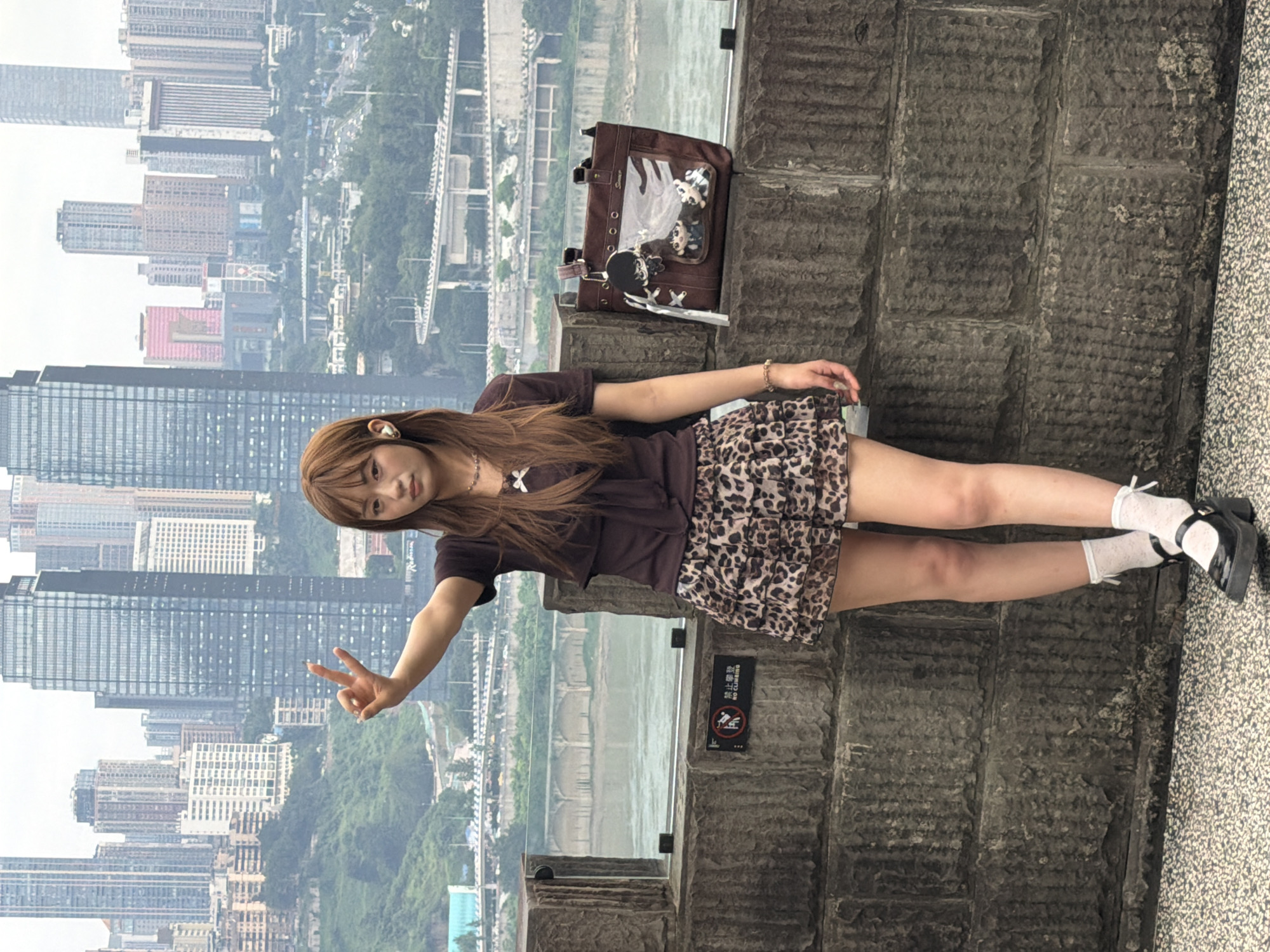 A young woman stands on a stone ledge overlooking a cityscape and river. She has long brown hair and is wearing a brown short-sleeved shirt, a leopard-print mini skirt, white ankle socks with ribbon ties, and black platform mary janes. She is holding her right hand up in a peace sign and has earbuds in her ears. A brown tote bag with cartoon charms hangs from the railing next to her. The background shows numerous tall buildings, a bridge, and a river, consistent with the Yuzhong District in Chongqing, China. The woman is positioned in the foreground, leaning slightly against a textured stone wall.