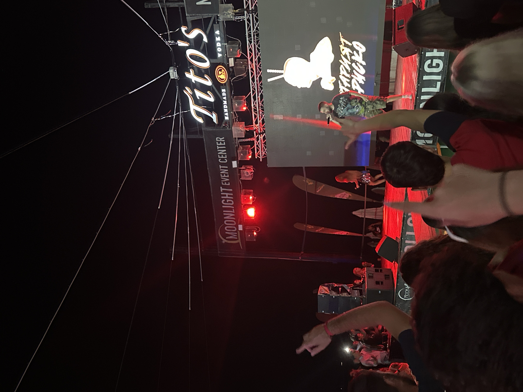 The image depicts an outdoor evening event in Varna, Bulgaria, possibly at the Moonlight Event Center, as indicated by signage. A stage is set up with a large LED screen displaying a graphic of a silhouette and text in Cyrillic, which appears to be "СТАРЫЙ РУЧКО". A male performer is on stage, singing into a microphone and wearing a patterned t-shirt and camouflage shorts. He is illuminated by stage lights. In the background, a female performer in a sparkly skirt can be seen. Above the stage, a prominent sign reads "Tito's HANDMADE VODKA". The foreground is filled with a crowd of people, many of whom are looking towards the stage. Some individuals in the crowd are raising their hands, suggesting engagement with the performance. The overall atmosphere appears to be a lively concert or festival.