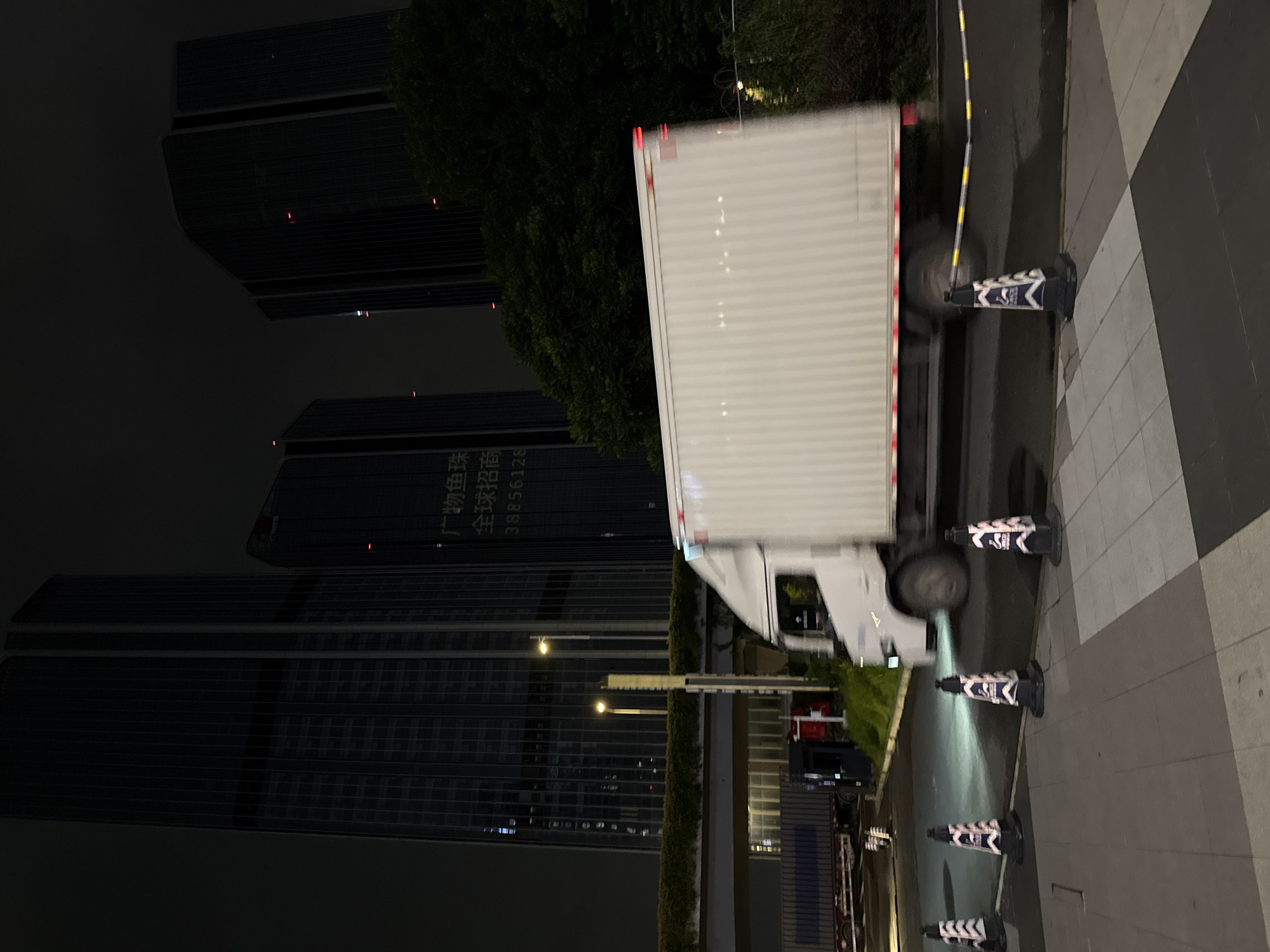 At night in Huangpu District, China, a white refrigerated truck is captured in motion, indicated by motion blur on its wheels and the background. The truck, a cube-shaped cargo vehicle, is positioned on a road, its rear end and side visible. The background features modern skyscrapers with illuminated windows, suggesting an urban environment. A dense cluster of dark green trees is situated between the buildings and the road. In the foreground, several traffic cones with reflective white and black stripes are placed on a paved area, marking a boundary or path. A street lamp with a yellow light is visible behind the cones, and a section of an elevated walkway or bridge with greenery running along its edge is also present. The overall scene depicts a commercial or industrial area at night, with active transportation and architectural elements.