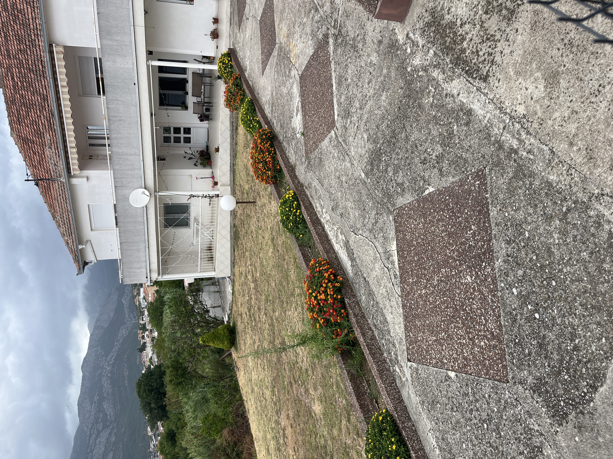 The image shows a white building with a red-tiled roof in a semi-rural setting. In the foreground, a concrete pathway with square inset sections leads towards the building. The pathway is flanked by a planter bed filled with colorful flowers, primarily orange and yellow. A dry, grassy lawn is visible to the left of the pathway. The building itself features balconies with white railings and an awning over the main entrance. In the background, a town with smaller buildings is nestled against a backdrop of misty mountains. The sky is overcast and gray.