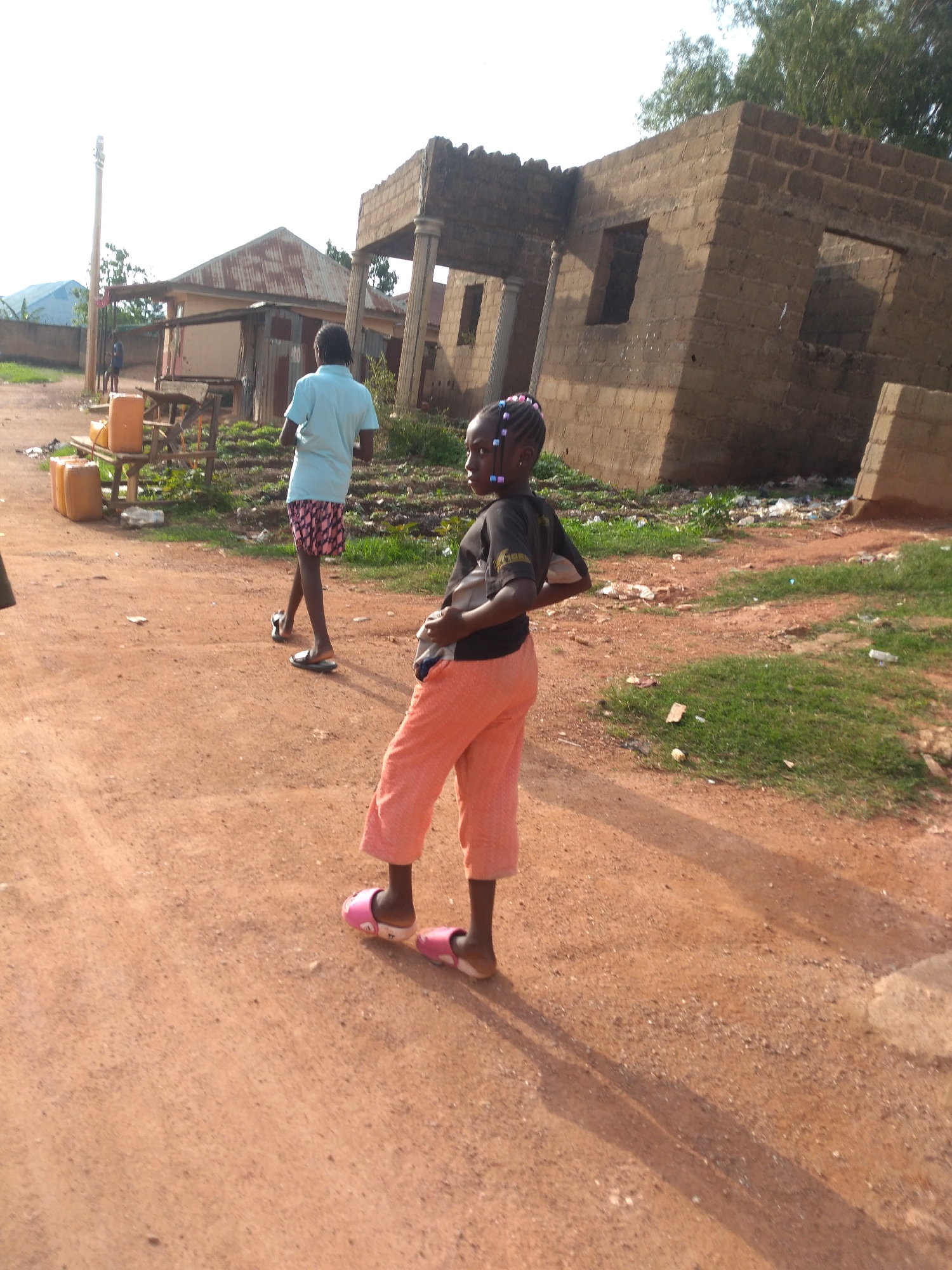 The image depicts a scene in Jos, Nigeria, with a focus on two individuals and their surroundings. In the foreground, a young girl is shown from the waist down, wearing orange pants and pink slip-on sandals. She is standing on a dirt road, facing away from the camera with her hands on her hips. To her left and further in the background, a person wearing a light blue shirt and patterned shorts stands on the same dirt road, also facing away from the camera.
The environment includes a partially constructed or dilapidated brick building in the right background, with visible windows and supporting columns. Adjacent to this structure is an area with green grass and scattered debris. To the left of the dirt road, there is a smaller building with a corrugated metal roof, and several yellow jerrycans are stacked on a wooden structure. A tall utility pole stands behind this building. The ground throughout the scene is predominantly reddish-brown dirt, indicative of the local soil composition in Jos. The lighting suggests it is daytime, with shadows cast on the ground.