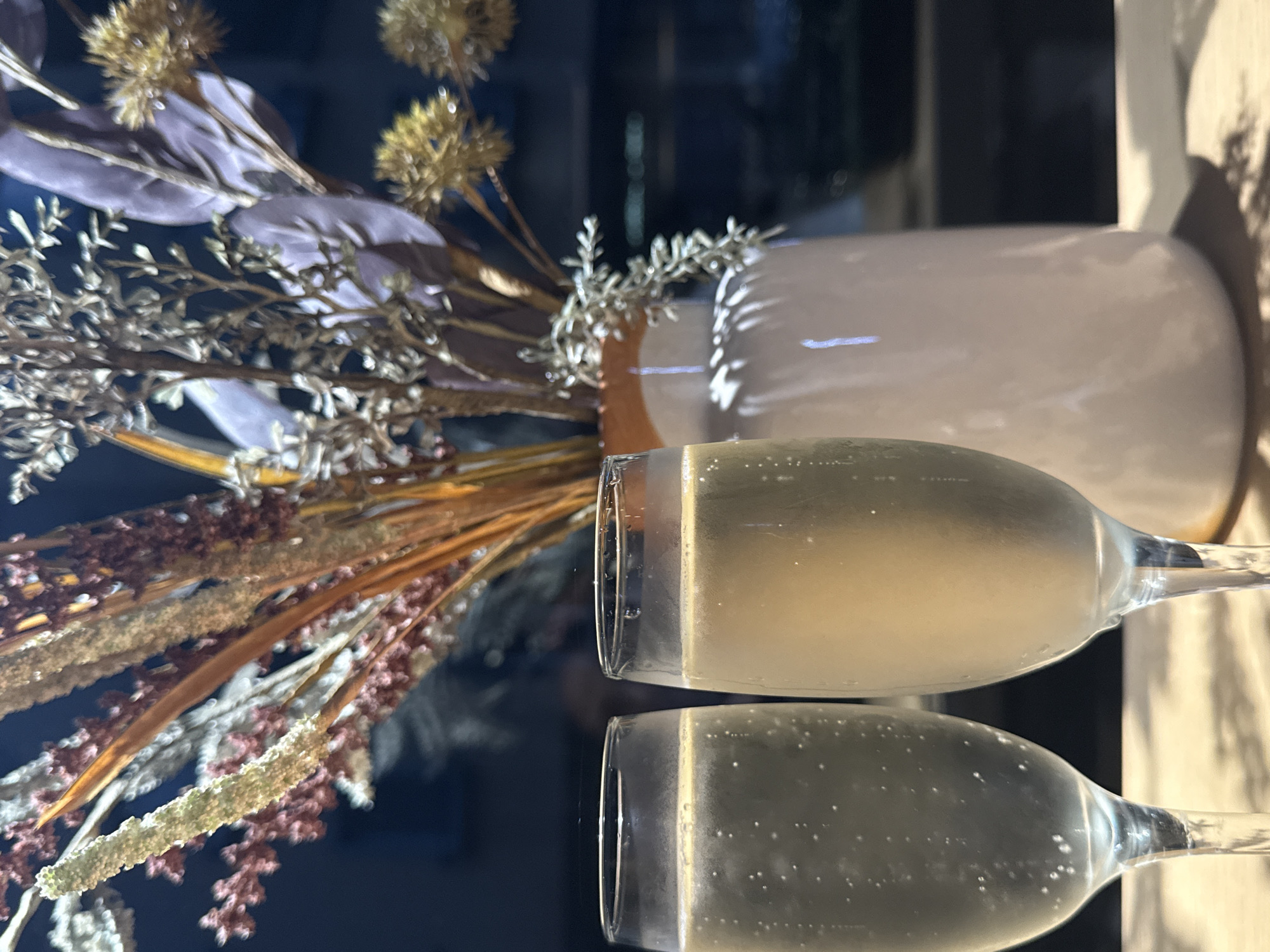 This image shows two champagne flutes filled with a pale yellow bubbly liquid, likely champagne or prosecco. The glasses are positioned vertically, one above the other, with the lower glass slightly closer to the viewer. The flutes are clear and show small bubbles rising within the liquid. The background is dark and out of focus, but appears to be a reflective surface, possibly a table or countertop.
To the left of the glasses, a bouquet of dried flowers and grasses is visible. The bouquet features a mix of textures and colors, including pale purple leaves, golden seed heads, and delicate brown and white grasses. The flowers are arranged in a somewhat wild and artistic manner. Behind the flowers and glasses, a pale pinkish-beige vase can be seen, its smooth surface reflecting some light. The lighting suggests it could be evening or a dimly lit indoor setting, with shadows casting across the scene, particularly on the right side of the vase.
The overall mood of the image is elegant and celebratory, hinting at a special occasion or a moment of quiet indulgence. There is no visible text in the image. Given the location context of Sofia, Bulgaria, this could be a scene set in a restaurant, home, or hotel room in the city. The reflective surface and the gentle lighting contribute to a sophisticated ambiance.
