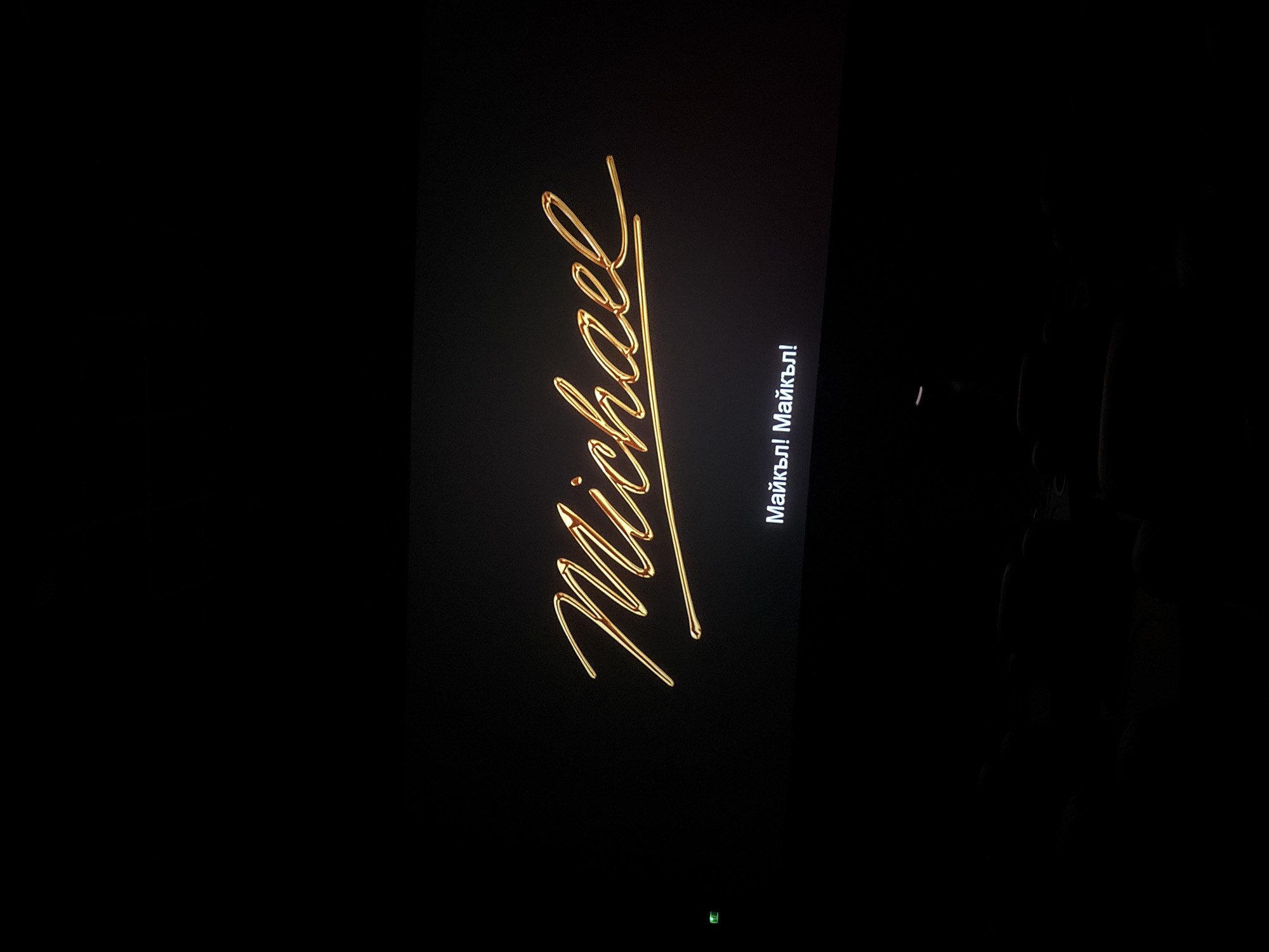 A screen displays the stylized golden cursive name "Michael" vertically, and beside it in what appears to be Cyrillic script, possibly Bulgarian, is the text "Майкъл! Майкъл!". The background is black. The location is unknown.