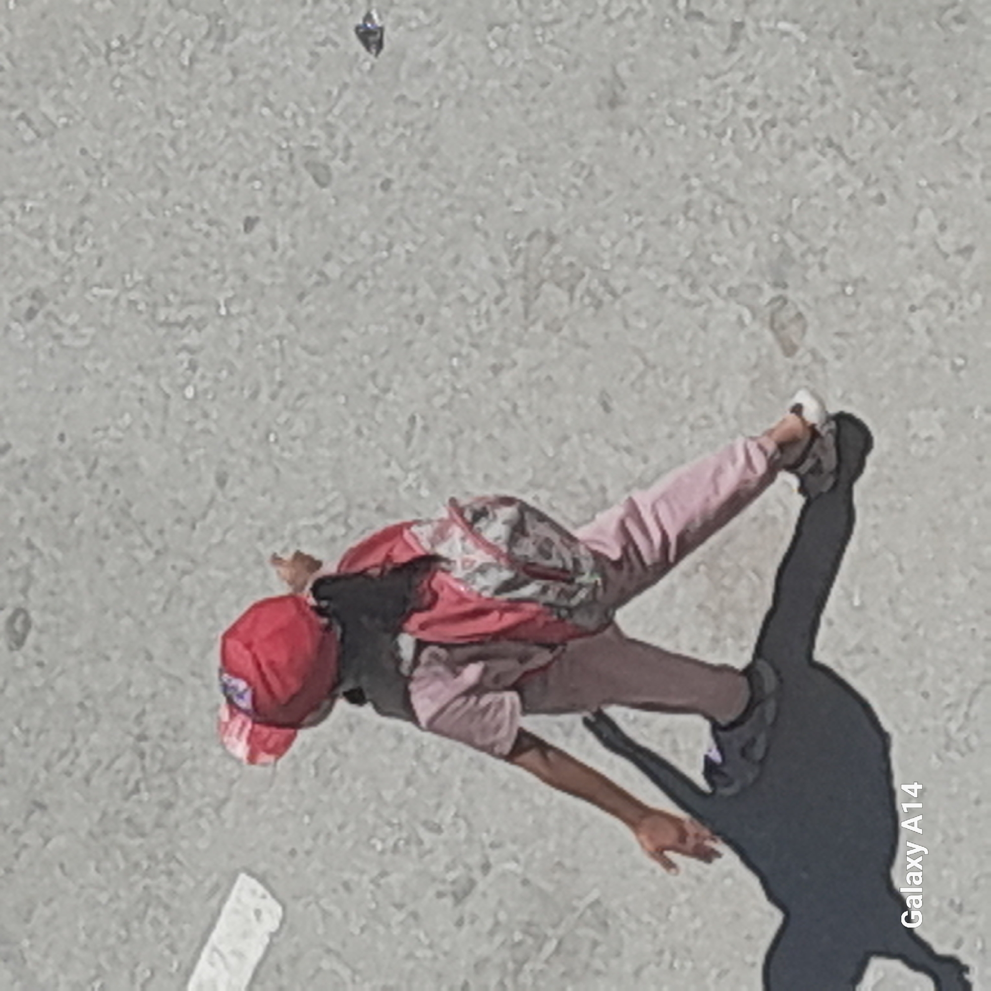 An overhead shot captures a child wearing a red hat and light pink clothing, walking on a paved surface. The child carries a backpack with a pink and white patterned design. A shadow of the child is cast on the ground. The surface appears to be asphalt or a similar coarse material, with some white markings visible. In the lower left corner, the text "Galaxy A14" is present. The scene is located in Khan Yunis, Palestinian Territories.