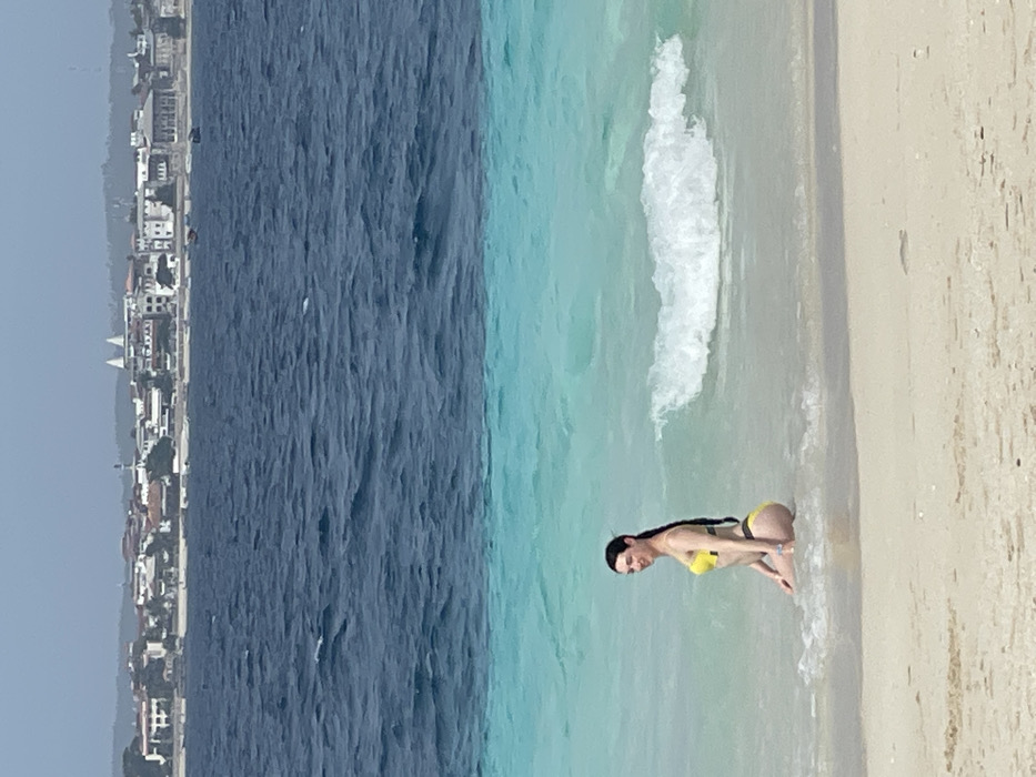 The image displays an outdoor coastal scene in Zanzibar, Tanzania. In the immediate foreground, a light-colored sand beach extends to the water's edge. A single adult female, with long dark hair, wearing a yellow bikini top and a dark bikini bottom, kneels in the shallow, clear turquoise water. She is positioned slightly angled to her right but looking towards the camera. A small white wave breaks approximately one meter to her right. The water deepens further from shore, transitioning to a darker blue and displaying ripples. The distant background reveals a coastal city skyline, characterized by numerous buildings of varying heights, some with red-tiled roofs and predominantly light-colored facades. Distinct white domed structures and minaret-like towers are visible within this urban landscape. Several small boats are discernible near the distant shoreline. The sky appears bright and overcast or brightly lit.