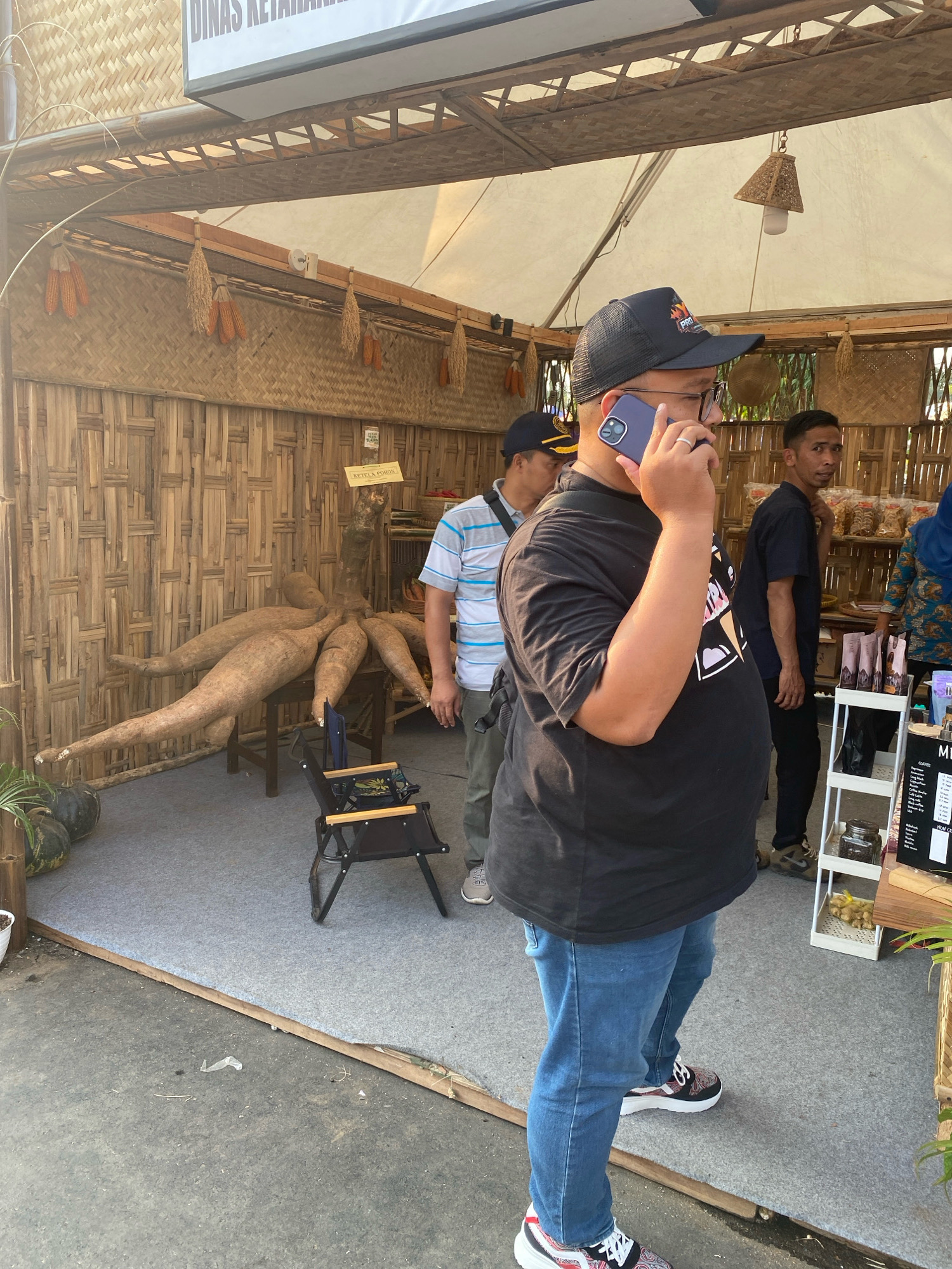 The image captures an outdoor market scene in the city of Nyamok, Indonesia, likely a local agricultural or craft fair, judging by the rustic bamboo stall construction. A man in a black t-shirt, blue jeans, and a black baseball cap is on his phone, positioned prominently in the foreground, obscuring parts of the stall behind him. To his left, another man wearing a striped t-shirt and dark pants appears to be observing the scene. Further back, a young man in a dark blue t-shirt and black pants looks towards the camera, possibly a vendor or attendee. Another individual in the background is partially visible, wearing a headscarf and a patterned top.
The stall itself is decorated with dried corn and rice stalks, and features a large, striking display of cassava roots. Various products are arranged on shelves, including what appear to be packaged snacks and coffee or tea related items. A small black folding chair sits on the grey carpeted floor within the stall. The overall environment suggests a sunny day, with shadows indicating ample natural light. The ground outside the stall is a rough, concrete-like surface.