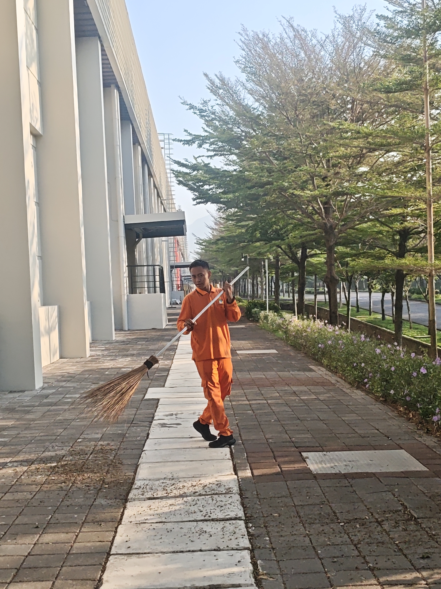 A male worker, wearing an orange uniform, is sweeping a paved walkway with a broom. He is facing the camera and appears to be in the process of sweeping debris. The walkway is lined with trees on the right and a modern building with white pillars on the left. The location is identified as Bae, Indonesia. The sky is clear and the lighting suggests it is daytime.