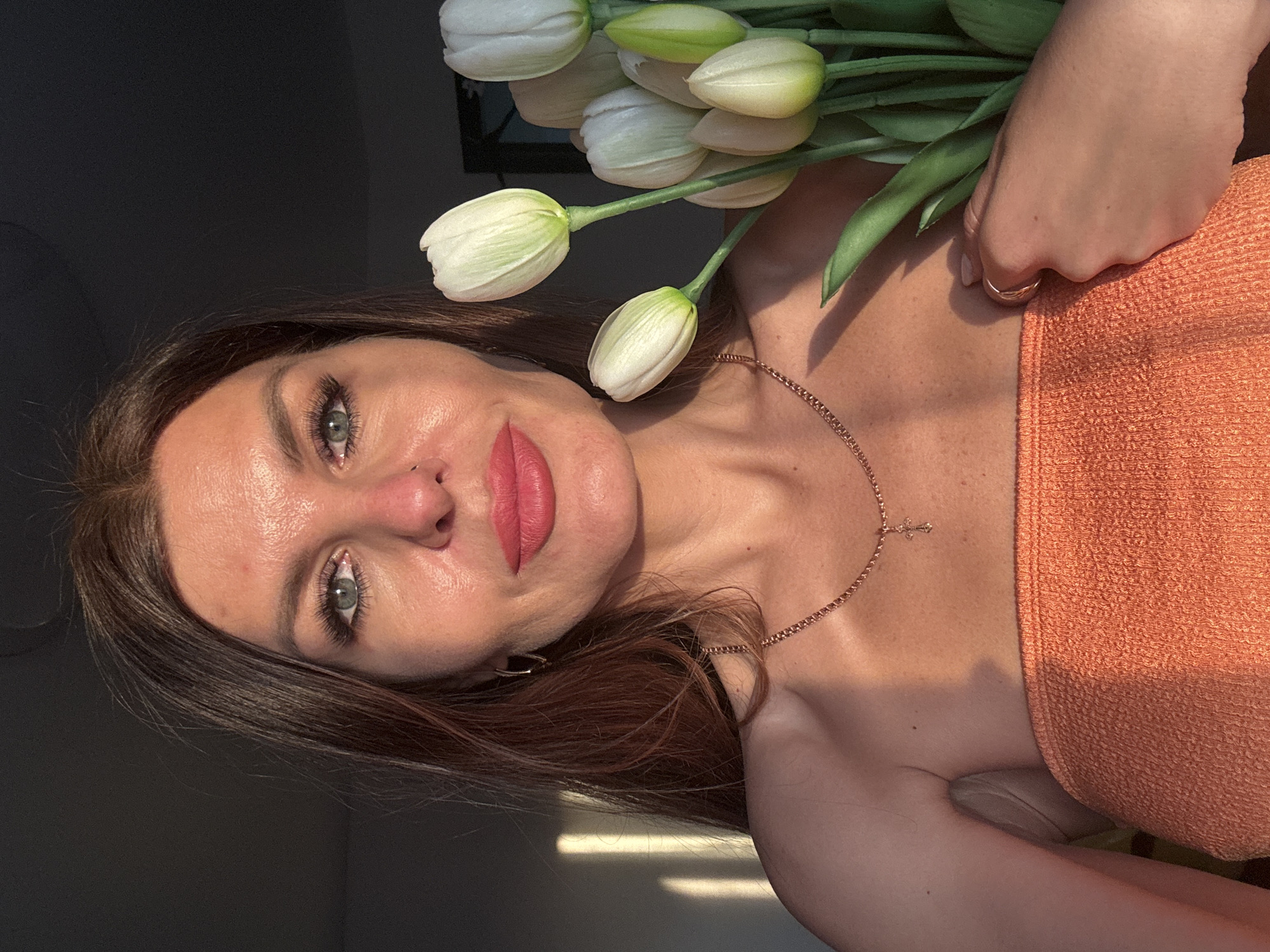 A woman is holding a bouquet of white tulips in her right hand. She is wearing a strapless, orange top. She has long, brown hair and light eyes. A thin, gold chain with a cross pendant is around her neck, and she wears a gold ring on her right hand. The background is dark. The location is Chaikovsky, Russia.