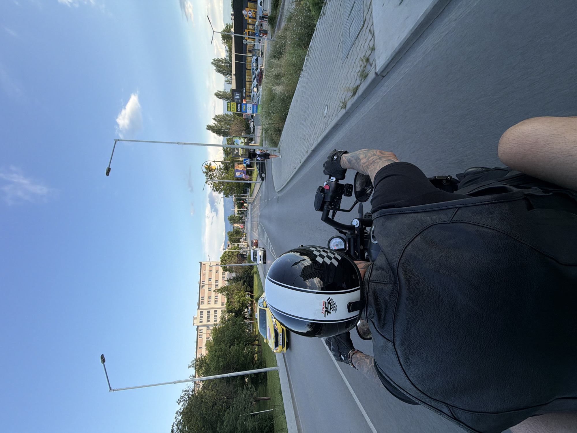 The image presents a first-person perspective from behind a person riding a motorcycle in an urban environment, presumably Plovdiv, Bulgaria. The rider, whose face is not visible, is wearing a black leather jacket and black gloves. Their forearms are heavily tattooed. A distinctive white helmet with a black checkered pattern on top and a black stripe along the center is worn, displaying "LOUIS 3S" text and a small checkered flag logo.
The motorcycle's handlebars, controls, and a dashboard with gauges are partially visible. The vehicle is on an asphalt road with white lane markings. In the immediate vicinity, a yellow taxi is visible ahead, followed by a white car, indicating traffic.
The setting includes various buildings. To the left, residential buildings with multiple windows line the street. To the right, a large commercial building or retail park features prominent signs for "BILLA", "IKEA", "TED", "JOY", "ОБУВКИ" (shoes), and "ДОМАШНИ ПОТРЕБИ" (household goods). A small wind turbine stands in the distance near this commercial area.
The scene is illuminated by daylight, suggesting a clear and sunny day, possibly late afternoon, with a bright blue sky and scattered white clouds. Trees, bushes, and grass are present along the roadside and pavement. Streetlights and a billboard are also visible. Distant mountains can be seen on the horizon. The visible Bulgarian text on commercial signs ("ОБУВКИ", "ДОМАШНИ ПОТРЕБИ") confirms the location context of Plovdiv, Bulgaria.