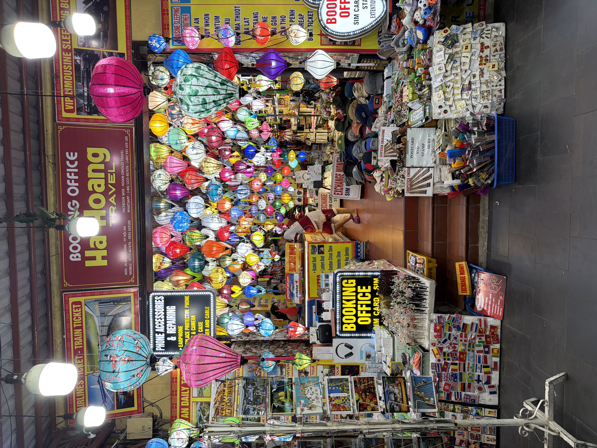 The image shows a street scene in Hội An Ward, Vietnam, characterized by a vibrant display of colorful lanterns and various shops. In the foreground, a market stall features a collection of small items, including postcards, flag stickers, and what appear to be souvenirs. A blue basket filled with various colored pens is also visible. Behind this stall, a stall with an "EXCHANGE MONEY" sign is present, with a woman and a child interacting with the counter.
Further back, the scene opens up to a more extensive display of goods, predominantly hanging lanterns of various sizes and colors, illuminating the area with a warm glow. Above the lanterns, shop signs are visible. One prominent sign reads "BOOKING OFFICE Hoang TRAVEL," with contact information and social media handles. Another sign advertises "AIRLINES TICKET - TRAIN TICKET." To the right, a sign indicates "VIP LIMOUSINE SLEEPING BUS."
The environment appears to be an outdoor marketplace or a street lined with shops, typical of tourist areas. The lighting suggests it could be evening or an indoor/covered market setting illuminated by artificial lights and the lanterns. The overall impression is one of a bustling commercial area, rich in local culture and offerings.