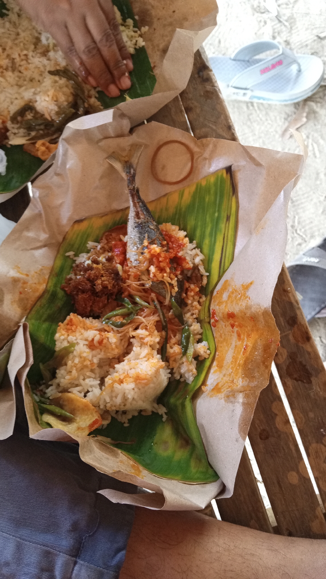 A close-up, overhead shot captures a meal served on banana leaves, wrapped in brown paper, and placed on a rustic wooden table. The meal consists of white rice, a whole fried fish, and a side of shredded meat or vegetables. A hand is visible in the upper left, reaching towards the rice. In the background, a pair of light blue flip-flops rests on a sandy or gravelly surface, suggesting an outdoor or beachside setting in Kebonbaru, Indonesia. The overall impression is of a casual, local dining experience.