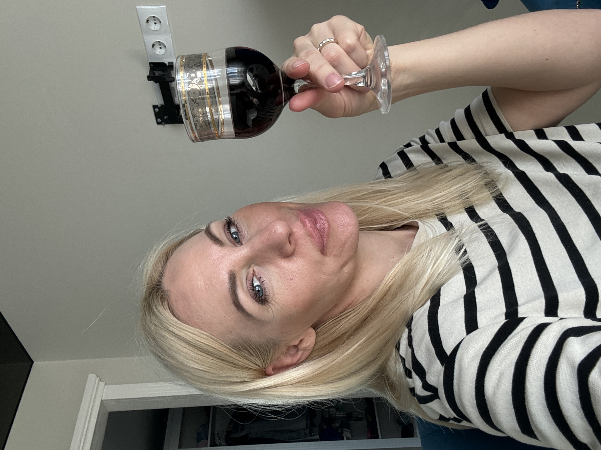 A woman with blonde hair, wearing a black and white striped shirt, holds a glass of red wine. She is indoors, and the background shows a wall with an electrical outlet and a closet. The scene appears to be in a residential setting.