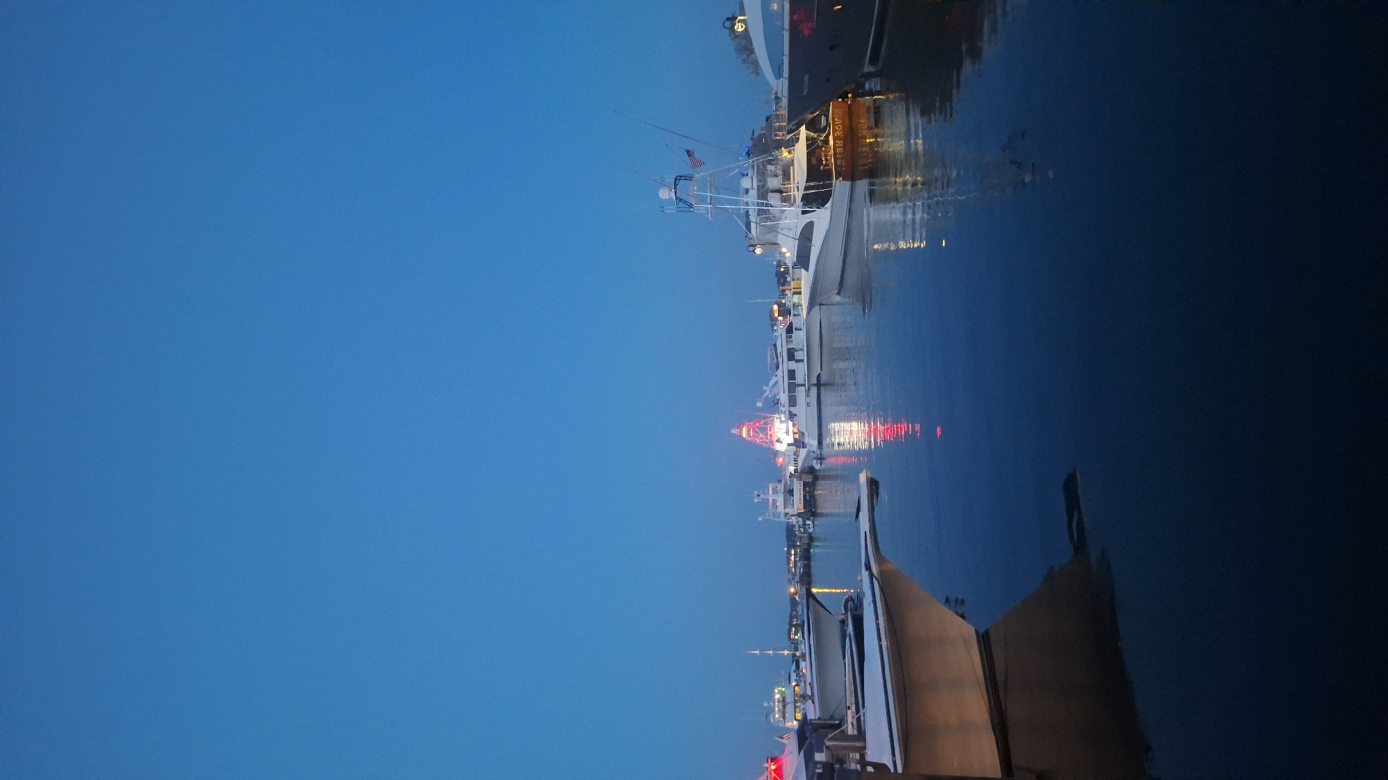 This is a vertical, low-angle shot of a harbor at dusk in Nantucket, Massachusetts, USA. The sky is a deep, clear blue. Several boats of various sizes are docked or anchored in the water, their reflections shimmering on the calm surface. In the background, a prominent red and white structure, likely a lighthouse or a similar landmark, is illuminated with red lights. The lights of the boats and the background structure are also reflected in the water. The foreground features the hull of a boat, partially submerged, creating a dark, abstract shape in the water. The overall atmosphere is tranquil and serene, with the fading light casting a soft glow over the scene.