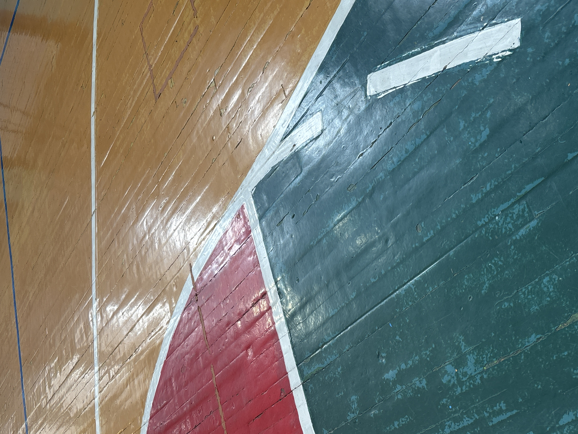 The image displays a close-up, high-angle view of a heavily worn wooden sports court floor, identified as being located in Tayshet, Russia. The surface exhibits extensive degradation, with widespread cracking, chipping, and peeling of paint and varnish across the floorboards. The court features a reddish-brown main area, delineated by a thin blue line in the upper background and faint, faded rectangular outlines suggesting prior markings. This reddish-brown section transitions into distinct red and dark green (teal-colored) painted zones, separated by worn white boundary lines. Both the red and dark green sections show significant paint loss and chipping, revealing the underlying wood grain. A prominent, elongated white marking is present on the dark green section, overlaying a broader, irregularly shaped white area. The floor's glossy, reflective patches indicate an indoor setting with overhead illumination. No individuals, objects other than the floor itself, or specific actions are depicted.