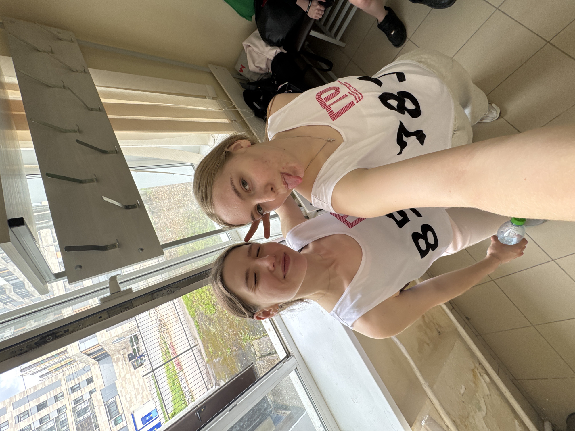 Two light-skinned young women are posing for a selfie indoors in Perm, Russia. The woman on top is sticking her tongue out and making a peace sign. Both women are wearing white tank tops with black lettering that reads "78" in a stylized font. The woman on the bottom is holding a plastic water bottle. They are standing near a window that offers a view of a building and outdoor greenery. Other objects visible include a clothes drying rack, clothes, and tile flooring.