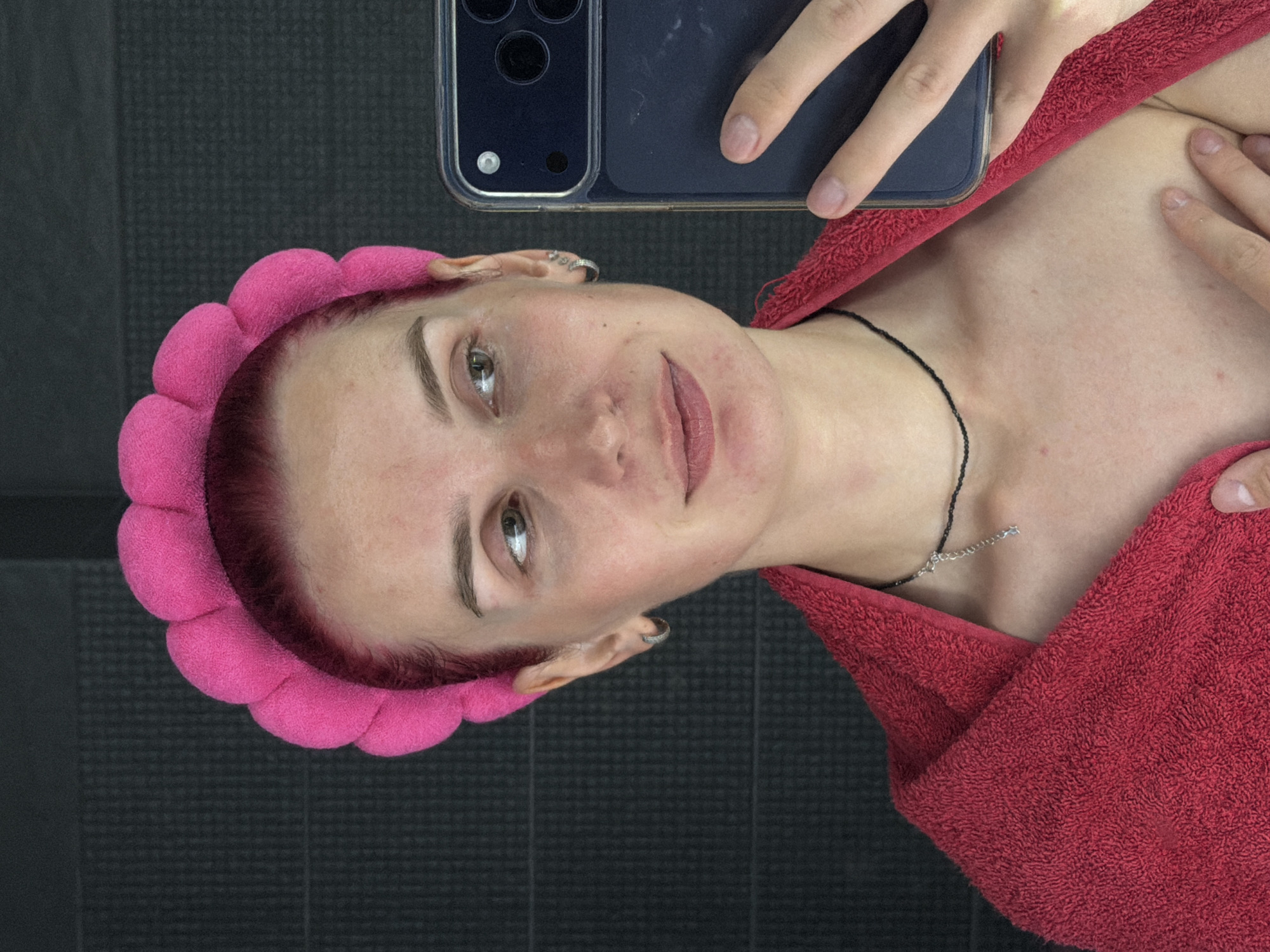 A selfie-style photograph captures a young woman from a top-down perspective, rotated 90 degrees clockwise. She appears to be lying down or reclining, looking upwards towards a smartphone she holds with her right hand. The phone, dark in color with a clear case and visible camera bump (resembling an iPhone), is positioned in the upper left of the frame, partially obscuring her head.
The woman has fair skin, with some minor redness or blemishes visible on her forehead, cheeks, and neck/chest area. Her hair, visible at the hairline, is dyed a reddish-pink hue and is held back by a bright pink, puffy, segmented headband. She wears a thin, dark (possibly black) necklace with a small silver rectangular pendant. A small silver hoop earring is visible in her left ear. Her lips are a muted pink or red, and she has a calm expression with a slight, closed-lip smile. Her eyes, with light-colored irises, are directed towards the camera.
She is draped in a textured, red terry cloth towel, which covers her shoulders and upper chest. The background consists of a dark, textured surface, possibly a floor or wall with a grid pattern. The image is taken indoors, and no specific time of day or weather can be determined. The scene suggests a personal moment, possibly during a self-care routine in a bathroom or similar setting in Chaikovsky, Russia. No visible text is present in the image.