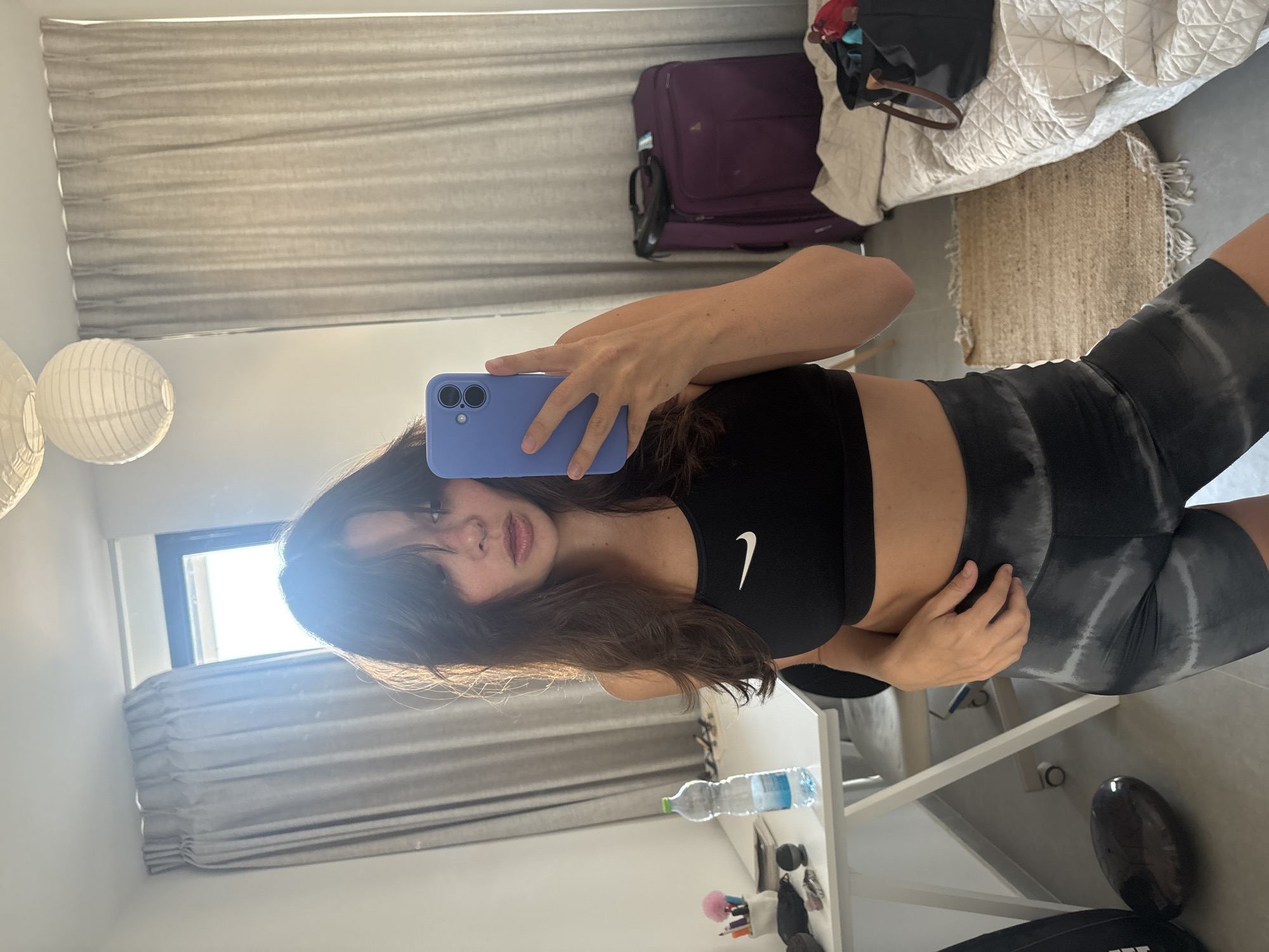 A young woman with long, dark, layered hair takes a selfie in a bedroom in Tel Aviv, Israel. She is wearing a black Nike sports bra and grey tie-dye biker shorts. She holds a blue iPhone in her right hand, which is angled to capture her reflection in a mirror. To her left, a white desk holds a water bottle, various pens and makeup brushes in a cup, and other small items. Behind her, a window with grey curtains is visible. To her right, a purple suitcase sits next to a bed with a quilted comforter. A woven rug lies on the floor between the bed and the woman. The room appears to be well-lit by natural light.