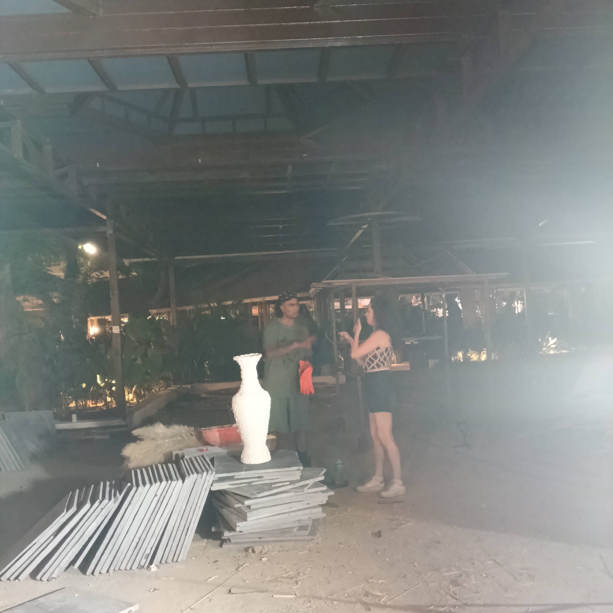 The image shows two individuals, one male and one female, standing in an outdoor area under a partially constructed wooden structure. The male is wearing a green t-shirt, dark pants, and a hat. He is holding orange gloves and appears to be in conversation with the female. The female is wearing a patterned crop top, a dark skirt, and light-colored sneakers. In the foreground, there are stacks of grey tiles and a white vase. Behind the figures are plants and structures that suggest an outdoor patio or garden setting. The image is heavily affected by light and shadows, with bright light sources causing glare and obscuring details. The location is inferred to be in Panaji, India, based on the background details.