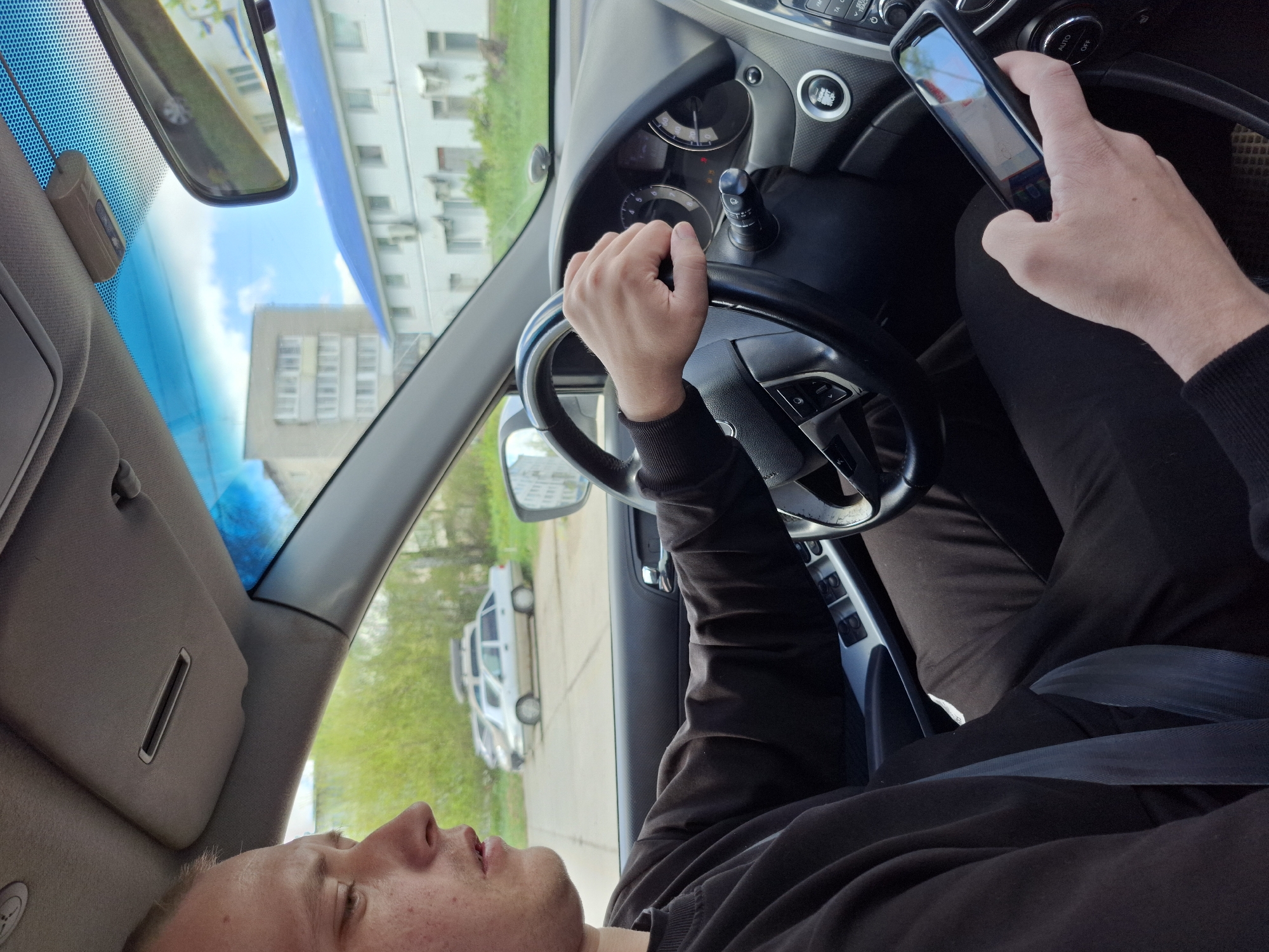 A person is driving a car. The individual has their left hand on the steering wheel and is holding a smartphone in their right hand. The driver is wearing a dark-colored long-sleeved shirt. The car is moving on a road with buildings and trees visible in the background. A white vehicle is visible outside the car window. The location is Chaikovsky, Russia.