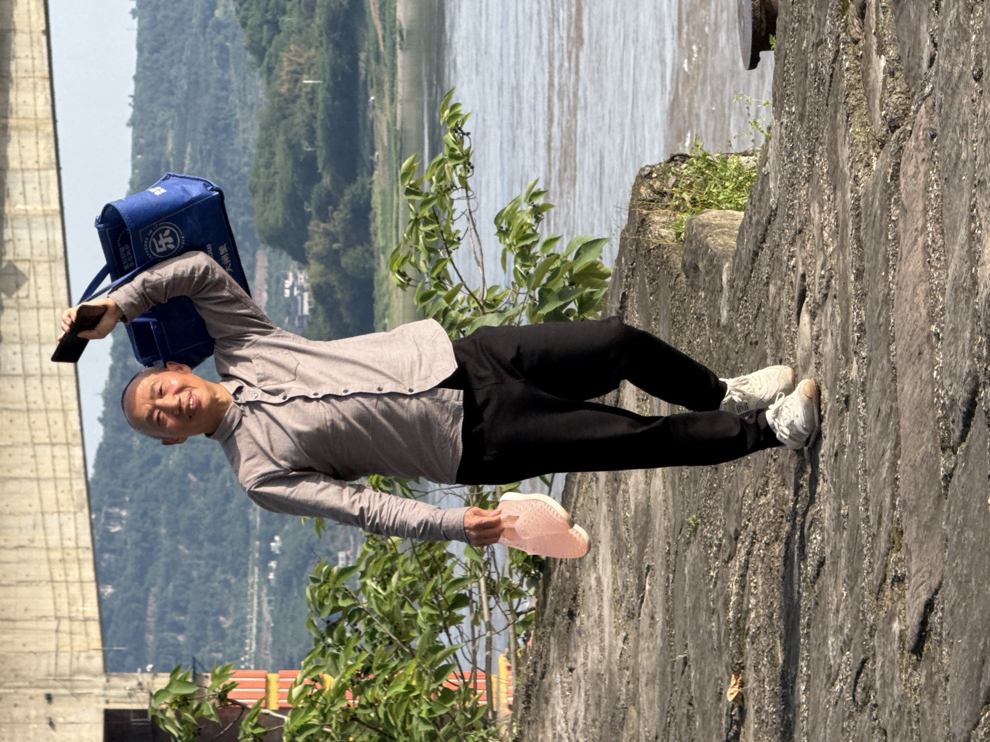 An Asian man stands on a concrete wall in Jiangjin, China. He holds a mobile phone in his left hand and a pink shoe in his right. The man has short, shaved hair and wears a gray button-down shirt and black pants. On his back is a blue bag. Behind him is a river, hills, and a partially-visible concrete structure. Green foliage is present near the man. The wall's texture is rough and gray.