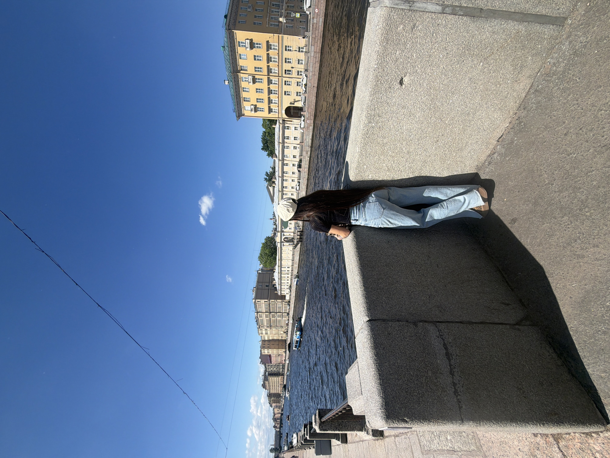The image depicts a woman standing on a stone embankment overlooking a wide canal in Saint Petersburg, Russia. The woman, seen from behind, has long dark hair and is wearing a black t-shirt, light blue wide-leg jeans, and a light-colored cap. She is leaning against a granite wall, facing the canal.
The canal is dark blue, with ripples on its surface indicating movement. Several boats are visible on the water, including a larger tour boat. The opposite bank of the canal is lined with buildings, typical of Saint Petersburg architecture, with multiple stories and light-colored facades. Some of these buildings have arched entrances and numerous windows. The sky is a clear, bright blue with a few scattered white clouds. In the foreground, the textured granite of the embankment wall is prominent, with a shadow cast by the wall. The scene appears to be bathed in bright sunlight, suggesting a warm, clear day. The overall impression is one of a scenic urban waterfront in Saint Petersburg.