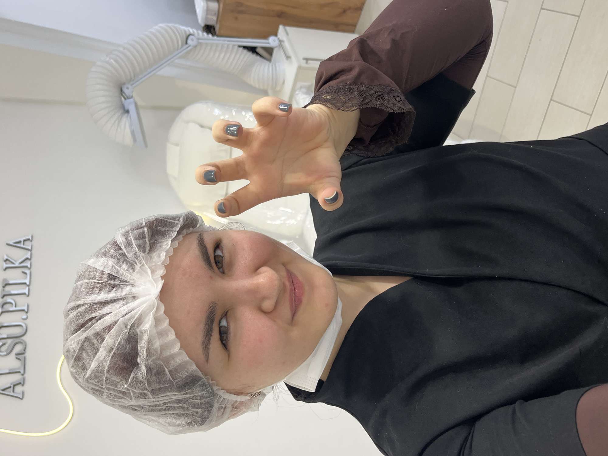 The image, oriented vertically, captures a close-up of a person indoors, likely in a salon or clinic setting. The person, who appears to be female, is positioned centrally and is looking directly at the camera with a slight smile. She has light-brown hair partially covered by a white disposable bouffant cap, commonly used in sterile or beauty environments. Her eyes are brown. A white disposable face mask is pulled down, resting under her chin. She is wearing a black top or smock, over which a dark brown long-sleeved shirt with lace detailing at the cuffs is visible on her right arm.
Her right hand is extended towards the camera, slightly out of focus, with fingers splayed in a playful 'claw' or 'grasping' gesture. Her fingernails are painted a glossy dark grey or charcoal color.
The background shows a brightly lit room with white walls and what appears to be white tiled flooring. To the upper left of the person, a flexible white hose, possibly part of a ventilation or extraction system often found in nail salons or medical settings, is visible. Above this, a wooden surface suggests a cabinet or shelf. On the left side of the image, running vertically along the wall, is a luminous sign with silver-colored letters that spell out "ALSUPILKA" in capital letters, with a thin yellow light strip tracing the bottom edge of the text. The overall lighting is bright and even.